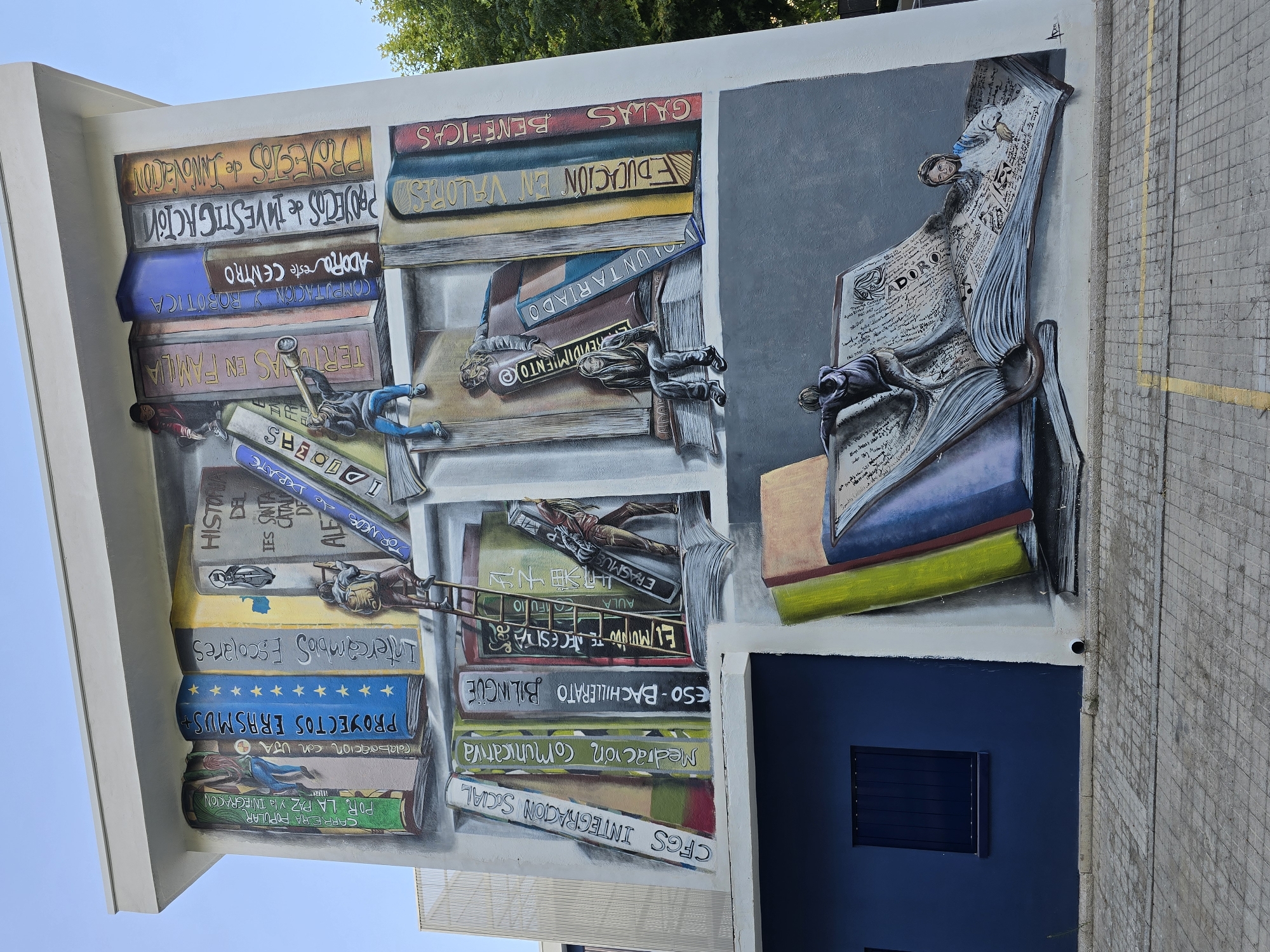 The image shows a mural painted on the side of a white building, depicting a large bookshelf filled with oversized books. The bookshelf and books are rendered in a realistic style, with various titles and images painted on the spines. Several figures, appearing to be students, are depicted interacting with the books in different ways. In the upper section, one figure climbs a ladder towards a book, while another looks through a telescope at a distant book. In the middle section, two figures are seated on a stack of books, one looking down and the other engaged with a telescope. In the lower section, two more figures are interacting with an open book, with one climbing on its pages and the other reclining within it. The mural is colorful and detailed, creating an illusion of depth and three-dimensionality. The building itself has a dark blue section on the left with a window and shutters. The ground in front of the building is paved with interlocking grey stones. Trees and a clear blue sky are visible in the background. The location is not specified in the image.