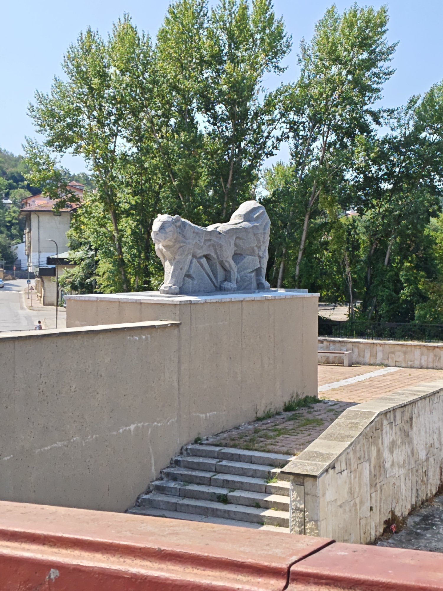 The image captures an outdoor scene with a prominent stone sculpture of a crouching feline, possibly a lion or a tiger, positioned on a raised platform. The sculpture is abstract and geometric in style. The platform is topped with a lighter-colored stone border. Below the platform is a textured concrete wall. To the left of the sculpture and wall, a street is visible with a few buildings and trees lining it. A lone figure is seen walking on the sidewalk. To the right of the platform, there is a paved area with a stone wall and what appears to be a bench. Stairs lead up from a lower level to the paved area. The background is filled with lush green trees, suggesting a warm, possibly summer day. This sculpture is located in the city of Lovech, Bulgaria, specifically at the entrance of the Vasil Levski High School.