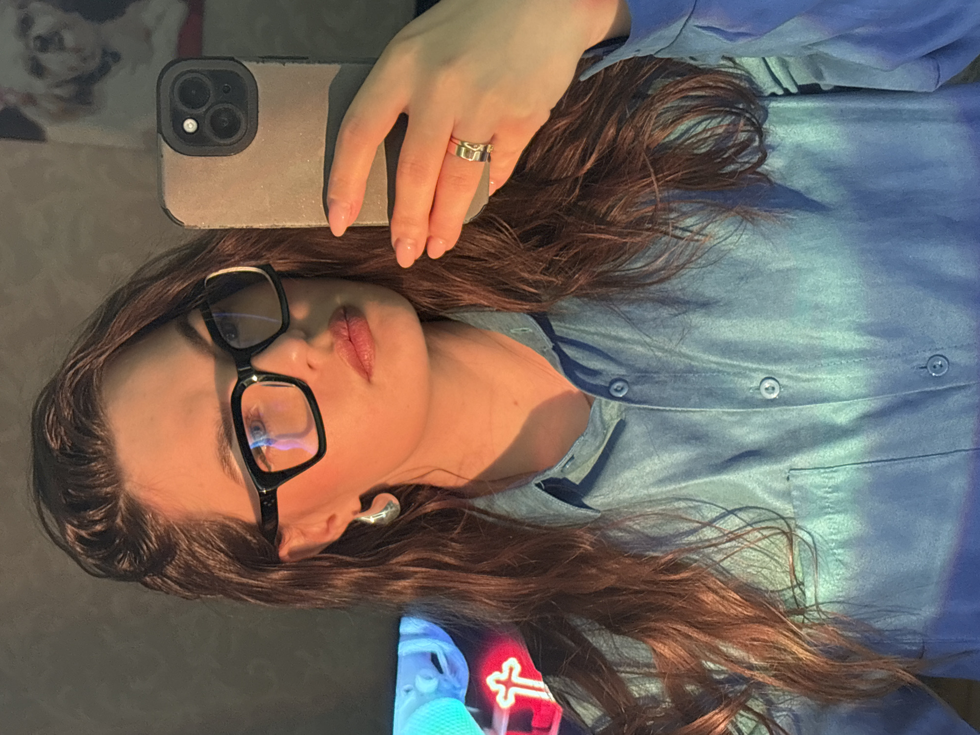 A person is reclining and holding a smartphone with three rear cameras. They are wearing glasses, a light blue button-down shirt, and a small earring in the visible ear. The person has long brown hair. There is a TV visible in the frame, near the bottom left.
There is not enough information to conclude that the image was taken in Chaikovsky, Russia.