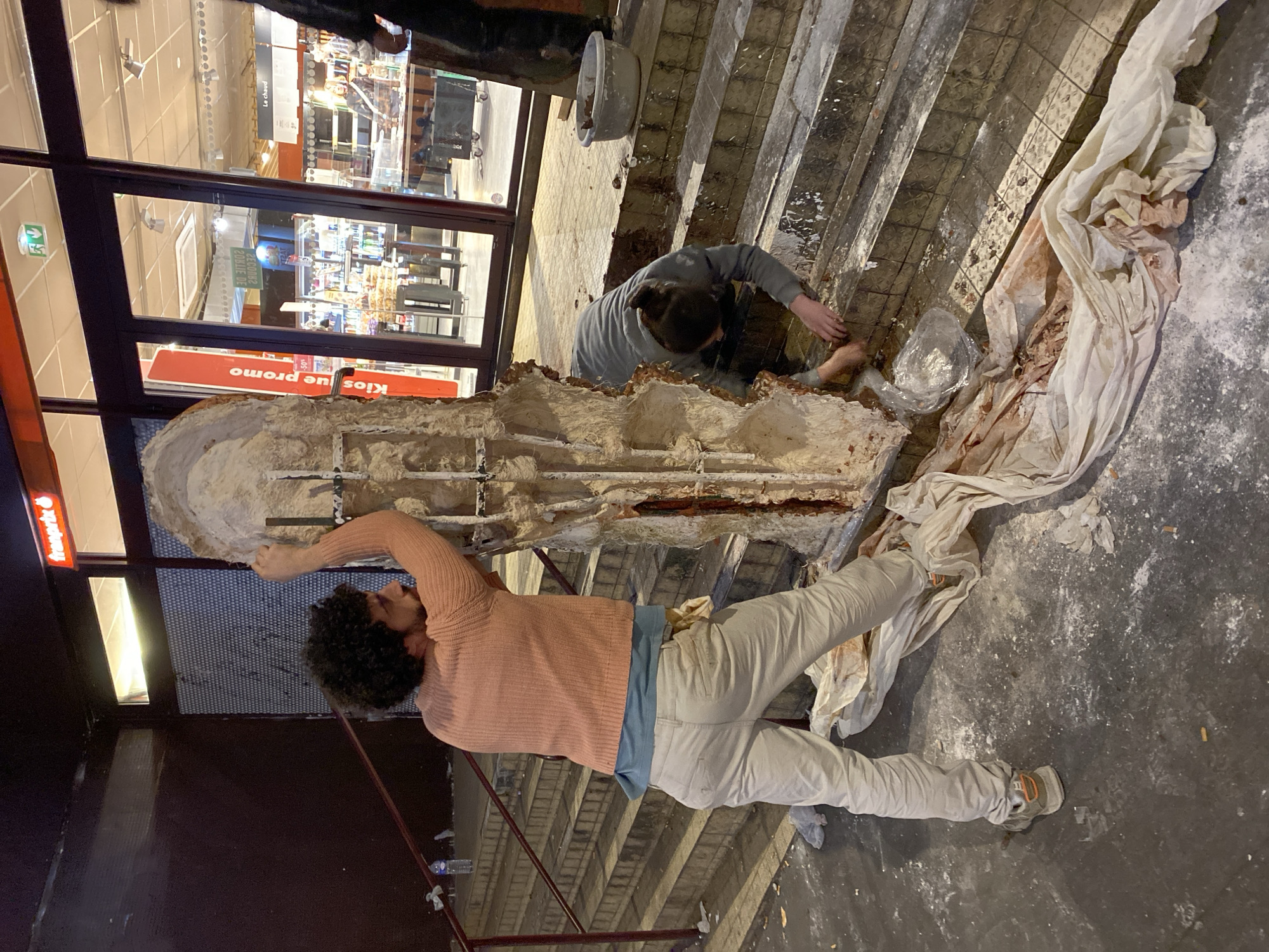 The scene is indoors. Two people are working on a column-like structure. One person is reaching out to the structure. He is wearing a pink sweater, a blue shirt, and light-colored pants. The other person is working on the base of the structure, wearing a grey sweater. The structure is partially covered with white cloth. They appear to be working in front of a shop. The reflection of the store name “fraprix” can be seen. The flooring and surroundings appear to be under construction. The location is in Paris, France.
