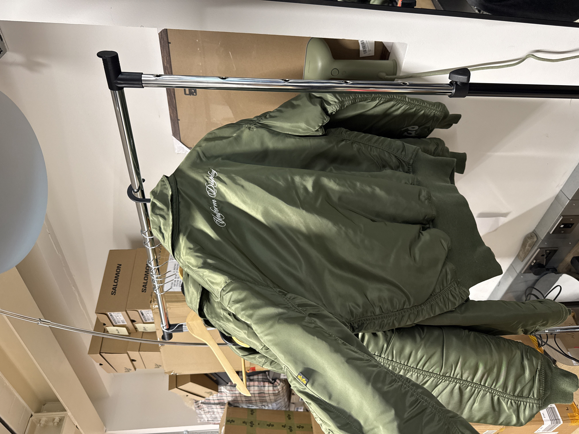 The image captures an indoor scene, likely a storage area or backroom in London, United Kingdom. The primary subject is an olive green bomber jacket, characteristic of an Alpha Industries MA-1 style, hanging on a silver metal clothes rack.
The jacket features white embroidered text on its back that reads "Maison Display" (likely "Maison Kitsuné Display") and a small yellow tag with the "ALPHA" logo is visible on its left sleeve pocket. The jacket has ribbed cuffs and hem, and appears somewhat padded. Several other garments or jackets are also hanging on the rack, partially obscured.
In the background, numerous cardboard boxes are stacked, prominently displaying the brand name "SALOMON" on their sides, suggesting they contain footwear or apparel. A light green, handheld garment steamer or similar device rests on a shelf or box behind the rack. The clothes rack itself has black plastic connectors and end caps. Part of a light grey spherical object, possibly a lamp or decorative item, is visible on the far left. The overall lighting is bright and artificial, consistent with an indoor commercial or storage space. No people are visible, and no specific activity is taking place, but the setup suggests storage, inventory, or preparation of items.