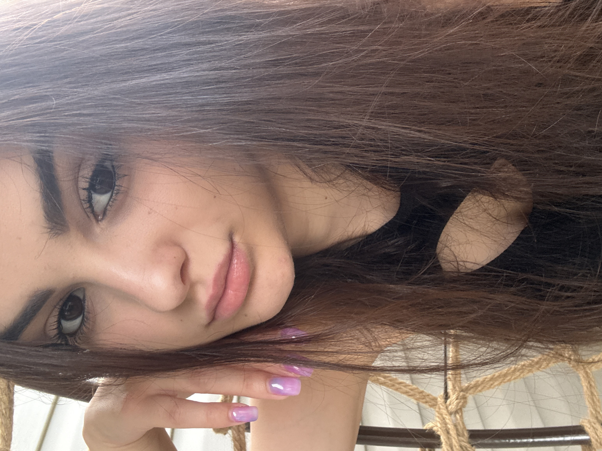 The media file is a close-up, low-angle, portrait-style photograph of a young woman. She has long, dark brown hair that frames her face and falls across the frame. Her face is turned to the side, with her left eye prominently visible and her right eye partially obscured by her hair. She has dark eyebrows, long eyelashes, and her skin appears fair with a subtle sheen. Her lips are slightly parted and appear to be colored with a light pink or nude lipstick. Her right hand is raised and resting near her face, with her fingers gently touching her cheek. Her fingernails are visible and painted with a gradient of purple and pink, suggesting a cosmetic detail. The background is out of focus, but appears to be a dimly lit indoor setting with elements of rope and a light-colored material, possibly indicating she is sitting in a hammock or a chair with rope accents. The overall mood of the photograph is intimate and serene, with the soft lighting and her relaxed pose contributing to this feeling. No text is visible in the image. The context provided indicates the location is Polena, Bulgaria.