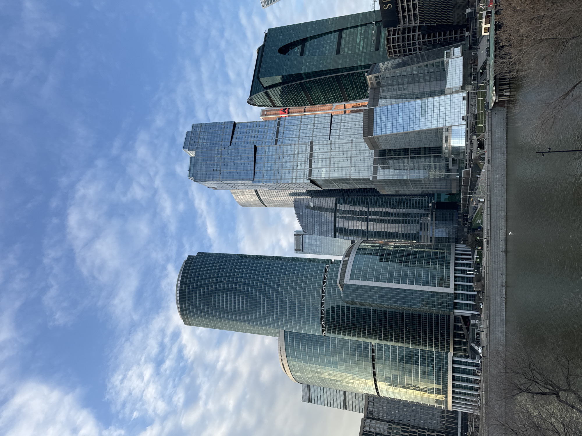 The image depicts the Moscow International Business Center, commonly known as Moscow City, located in Moscow, Russia, not Khimki. The scene presents a dense cluster of modern glass and steel skyscrapers of varying heights and architectural designs, situated along the Moskva River.
Prominent structures include the curved, multi-tiered glass facades of what appears to be part of the Federation Tower complex on the left, a series of rectangular and faceted glass towers in the center, and the distinctively twisting Evolution Tower on the right. The Evolution Tower features a prominent arched opening in its lower section. Reflections of the partially cloudy blue sky are visible across the numerous building facades.
In the foreground, the dark-colored waters of the Moskva River flow horizontally. A concrete embankment with a retaining wall lines the riverbank. Above the embankment, a multi-lane roadway is visible, with several light-colored vehicles in motion or parked. Pedestrians are discernible on sidewalks and staircases leading down to the river level. The immediate lower foreground contains bare-branched trees and shrubs, indicative of a non-foliage season. A single white bird is present on the river's surface near the right bank. On the far right, adjacent to the Evolution Tower, a partially constructed building with exposed structural framework and a crane arm is visible. The sky above is largely blue with scattered, wispy white clouds.