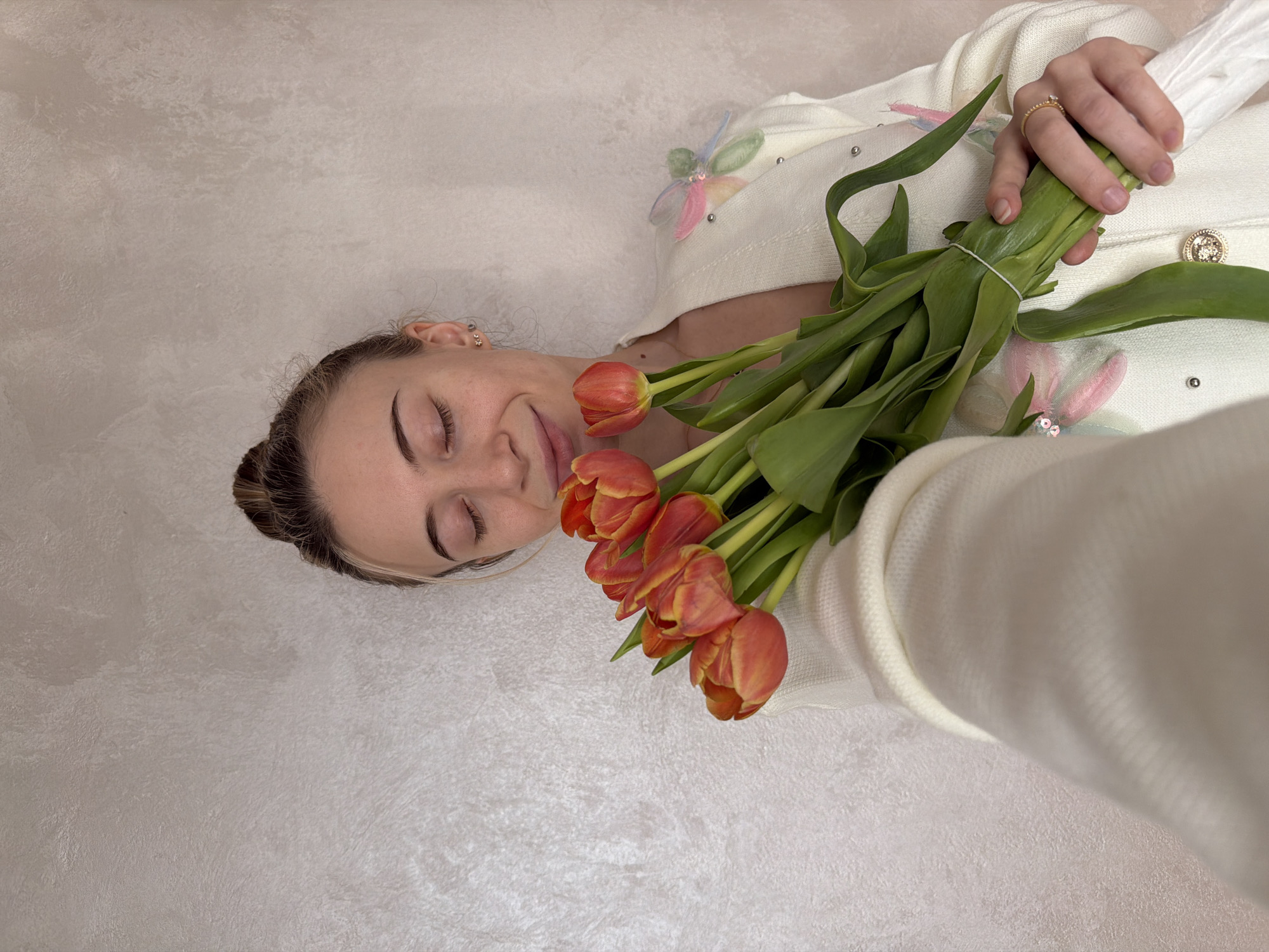A woman with fair skin and her eyes closed is lying down, holding a bouquet of orange tulips. Her hair is pulled back, and she is wearing a white cardigan with decorative flower details. She has a ring on her finger. The background appears to be a textured, light-colored wall. The location is unspecified, and it is unknown whether the scene is in the city of Plovdiv, Bulgaria.