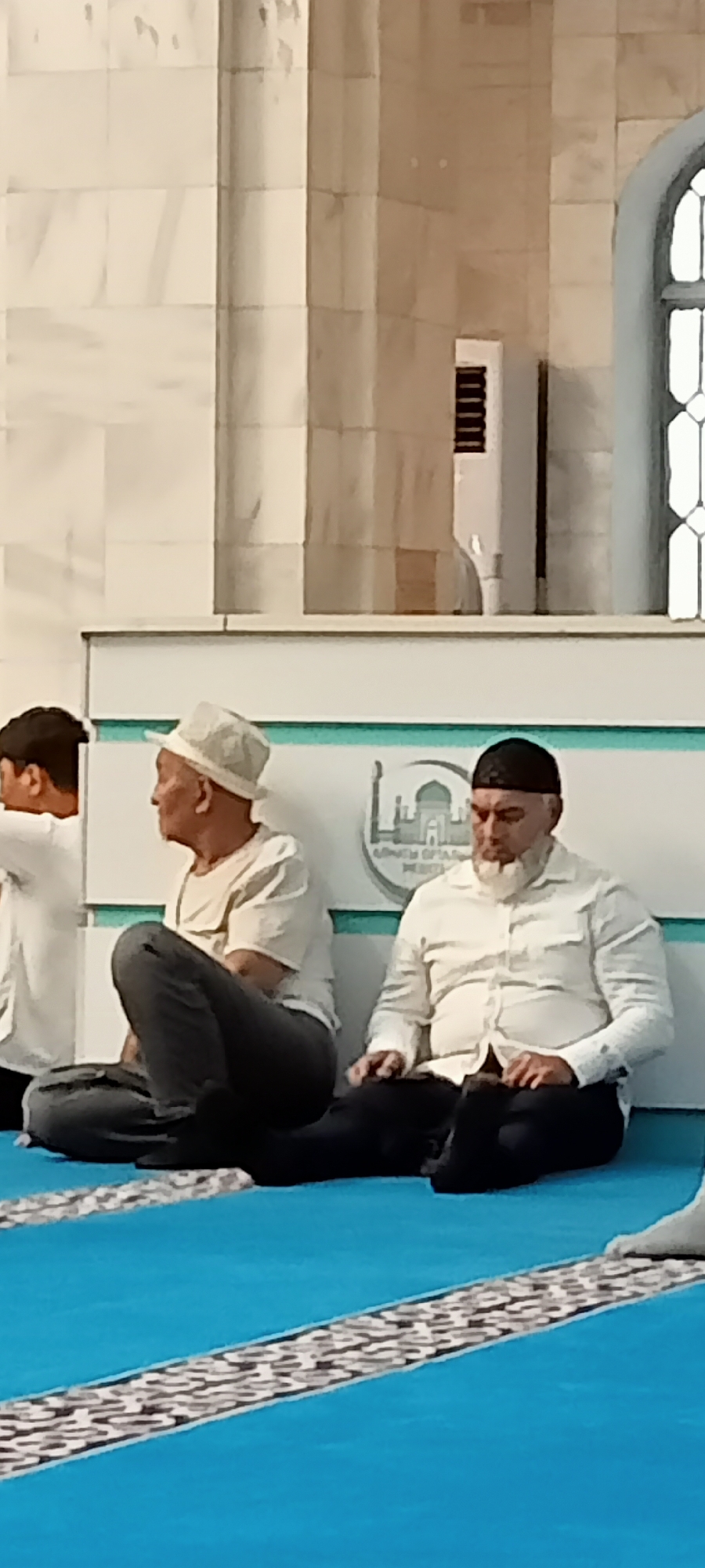 The image depicts an interior scene, likely a mosque, featuring several individuals in prayer or contemplation. The floor is covered in bright blue carpet with decorative white and black striped borders. In the foreground, two adult males are seated on the floor, facing left. The male on the right wears a white button-down shirt, black pants, and a black cap. He has a white beard. The male to his left wears a light-colored collared shirt, dark pants, and a light-colored fedora-style hat. He is seated with his legs crossed. To the far left, a young person, likely male, wearing a white t-shirt, is visible from the shoulders up, with dark hair. The background shows a wall with marble-like paneling and an arched window on the right. A white ventilation unit is mounted on the wall. A circular emblem or logo featuring a building with domes and minarets is visible on the wall behind the seated men, with text below it in an unfamiliar script, suggesting a location in Almaty, Kazakhstan, potentially related to a religious institution. The lighting appears to be indoor, with no strong directional sources, resulting in soft shadows.