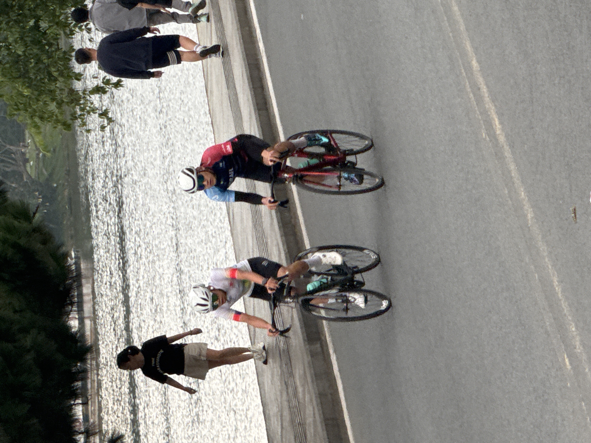 In Da Lat, Vietnam, two cyclists are engaged in a race on a paved road alongside a body of water. The cyclist on the left wears a white and black cycling kit and a white helmet, while the cyclist on the right is dressed in a red and blue cycling kit and a white helmet. Both are focused on the race, with the cyclist on the right appearing to be slightly ahead. Behind them, a woman in a black t-shirt and tan shorts walks along the side of the road. Further in the background, two men walk along the paved pathway. The environment is characterized by a large, shimmering body of water and lush green trees. The road is grey asphalt with faint white markings, and the pathway beside it is also paved. The scene captures a moment of athletic activity within a scenic natural setting.