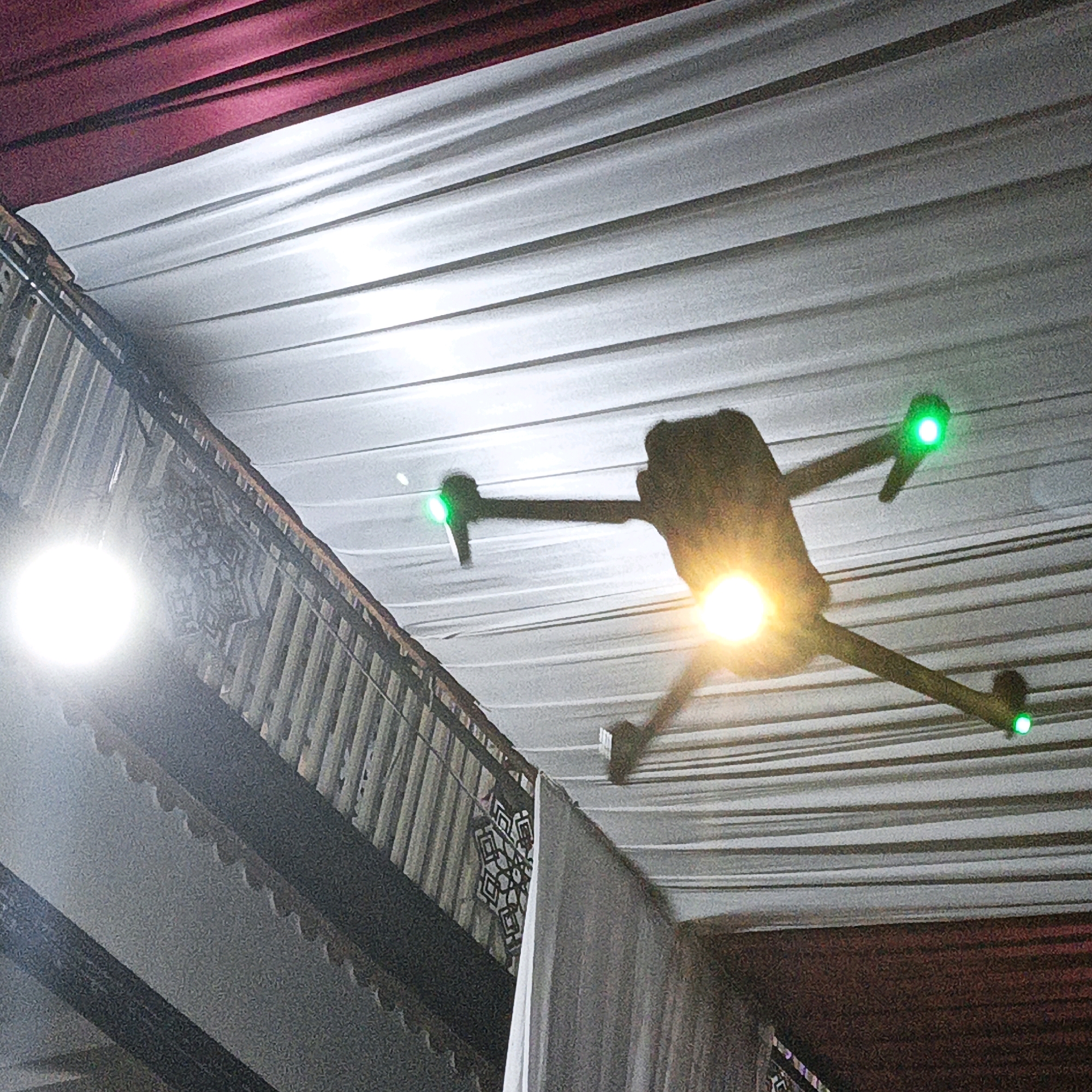 A drone is captured in mid-flight indoors, likely at an event venue in Kalitorong, Indonesia. The drone is silhouetted against a white draped ceiling, with red drapery visible at the top of the frame. The drone has three visible green lights, one on each of two arms and one on the body, and a bright yellow light emanating from its underside, presumably for illumination. The drone's shadow is cast on the ceiling. To the left, a bright white light source is visible, possibly a spotlight. Below and to the left of the drone, a decorative balcony railing with intricate patterns can be seen, along with a white curtain hanging down. The overall impression is of a dimly lit indoor space decorated for an occasion, with the drone actively recording or providing lighting.