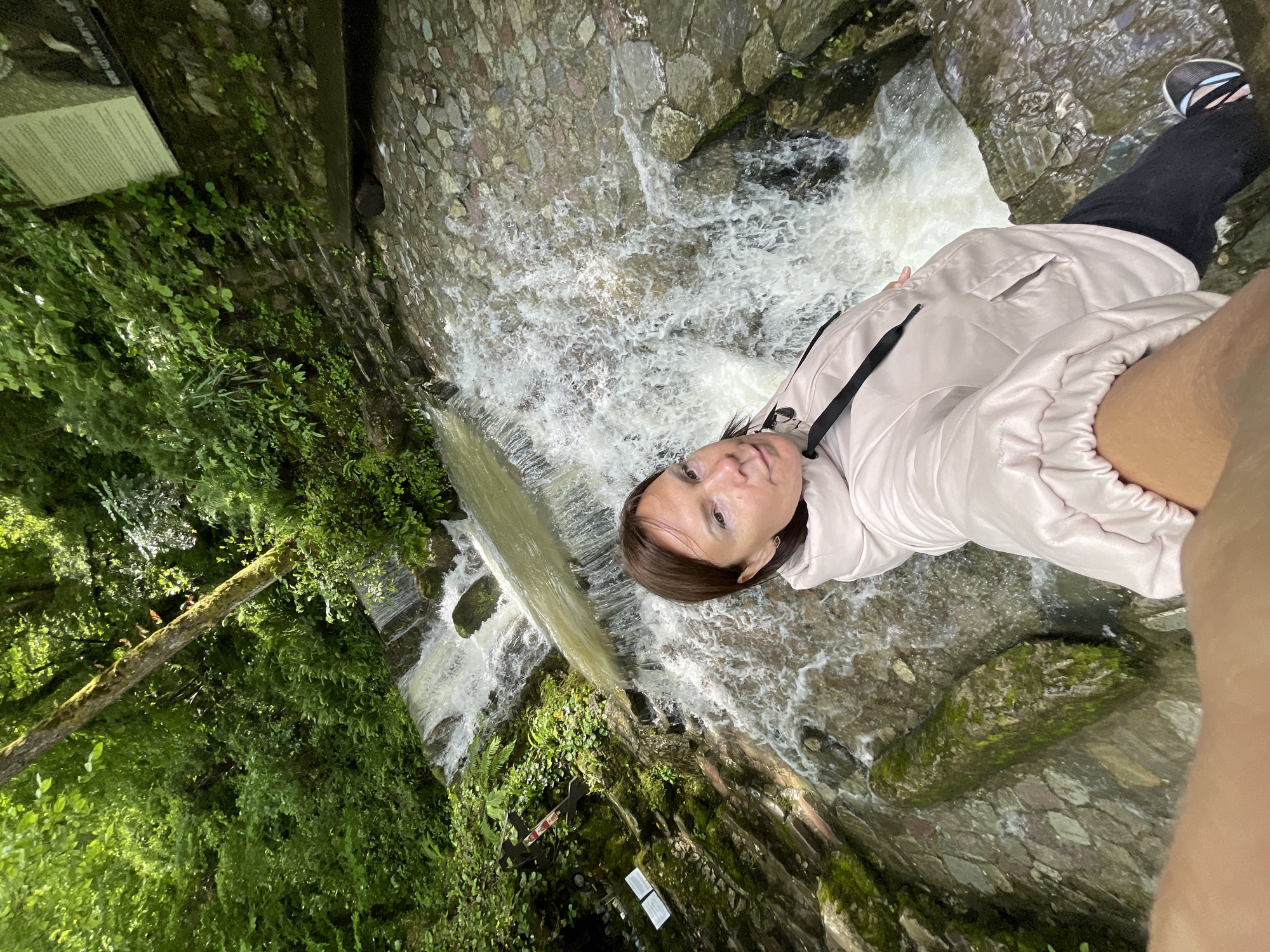 A woman is standing near a waterfall in Solokh-Aul, Russia. She is wearing a light pink jacket and dark pants, with black and blue sneakers. The waterfall is cascading over a rocky, stone-lined channel surrounded by lush greenery and moss-covered rocks. A sign is visible on the left side of the image, among the foliage. The picture appears to be a selfie taken at an angle.