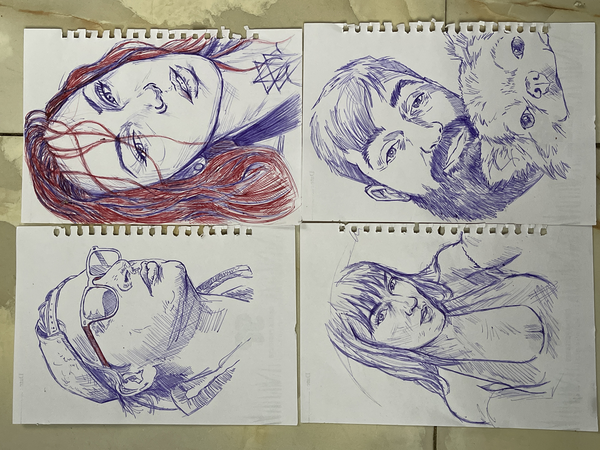 The image displays four pen-and-ink portraits on torn notebook pages, arranged in a 2x2 grid. The pages are laid out on a tiled floor, possibly in Abuja, Nigeria, indicated by the subtle presence of patterned tiles.
The top-left portrait is of a person wearing a baseball cap and sunglasses, depicted in profile facing right. Their head and shoulders are visible. The drawing utilizes blue ink with hatching for shading.
The top-right portrait is of a woman with reddish-brown hair, her face turned slightly towards the viewer. Red ink is used for her hair and some facial details, while blue ink is used for the rest of the drawing, including her shoulders and a star-like tattoo on her neck.
The bottom-left portrait features a woman with bangs and long hair, her gaze directed towards the viewer. Her upper body is shown, wearing a low-cut top. The drawing is done in blue ink.
The bottom-right portrait shows a man with a beard and a dog. The man's head is close to the dog's, and they are both looking towards the viewer. The drawing is executed in blue ink.
The torn edges of the notebook pages and the surface they are placed on suggest an informal setting.