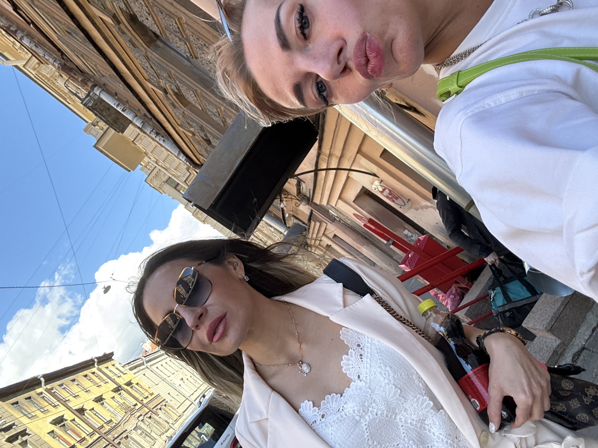 Two women are taking a selfie on a street in Saint Petersburg, Russia. The woman on the left is wearing sunglasses, a white lace top, and a light pink blazer. She is holding a bottle of dark liquid. The woman on the right is puckering her lips and has short blonde hair. She is wearing a white t-shirt and a green strap from a bag is visible. They are standing in front of buildings with ornate architecture, and the sky above is blue with some clouds. The street appears to be cobblestone.