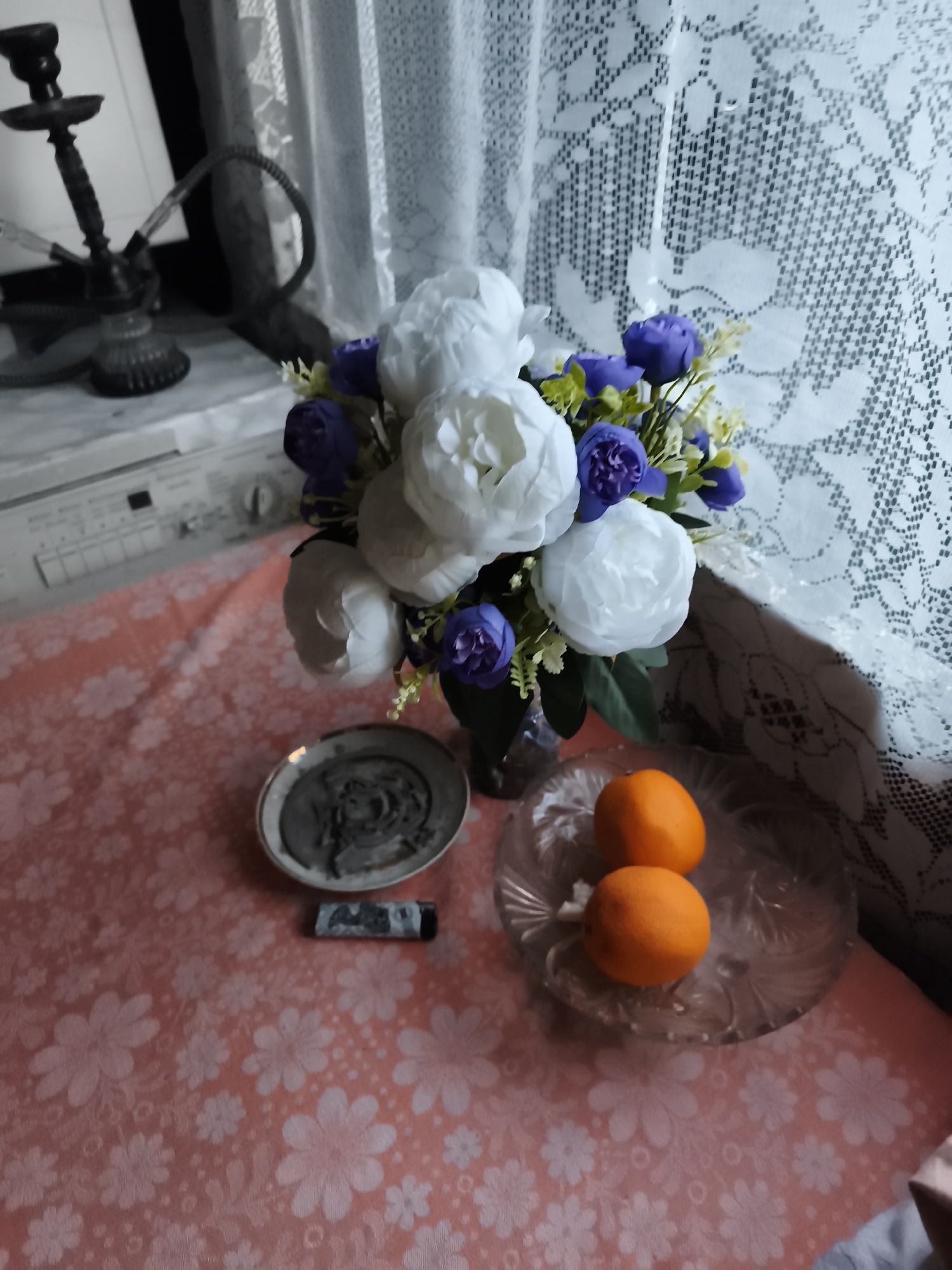 The image captures a still life arrangement on a table covered with a pink tablecloth patterned with white flowers. In the foreground, two oranges are placed on a clear, decorative glass plate. To the left of the oranges, a small, round dish contains what appears to be dark ash, possibly from incense or smoking. Beside this dish lies a black lighter.
Dominating the center of the frame is a bouquet of artificial flowers in a dark vase. The bouquet features large, white peonies and smaller, purple flowers with green foliage.
In the background, a white lace curtain with an intricate pattern is visible, suggesting an indoor setting, possibly near a window. To the left of the curtain and bouquet, part of a hookah pipe is visible, made of dark material with a coiled hose. Further left, a countertop or surface is visible, and behind it, a white appliance, likely a washing machine, can be seen.
The lighting suggests it could be daytime, with diffused light coming from the direction of the curtain. The overall scene is a collection of various objects arranged on a table, creating a somewhat eclectic still life. There is no visible text in the image. The location context provided is Daugavpils, Latvia.