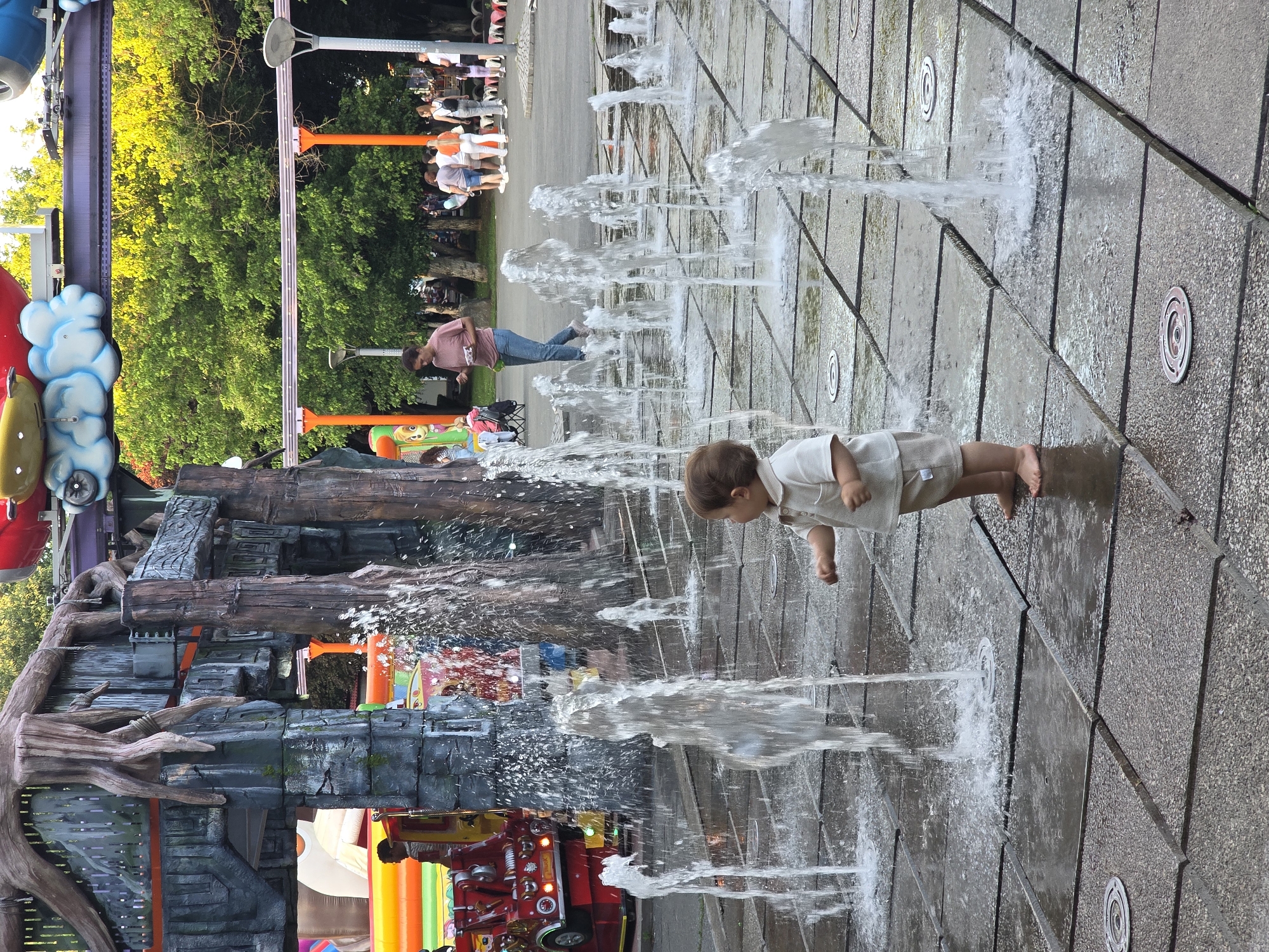A young child, approximately 2-3 years old, with brown hair, stands barefoot on a wet, stone-tiled surface in an outdoor amusement park. The child wears a light-colored polo shirt and beige shorts, with their right hand extended towards a water fountain. Several water fountains are active, spraying water upwards and creating a wet environment. In the background, various amusement park attractions are visible, including a large, tree-like structure with mossy accents and a red fire truck-themed ride. Behind this, a monorail track with orange supports runs through lush green trees. Several adults are also present in the background, walking and observing the attractions. The scene appears to be set during the daytime, with ample natural light. This amusement park is located in Bucharest, Romania, likely within the capital city's urban landscape.