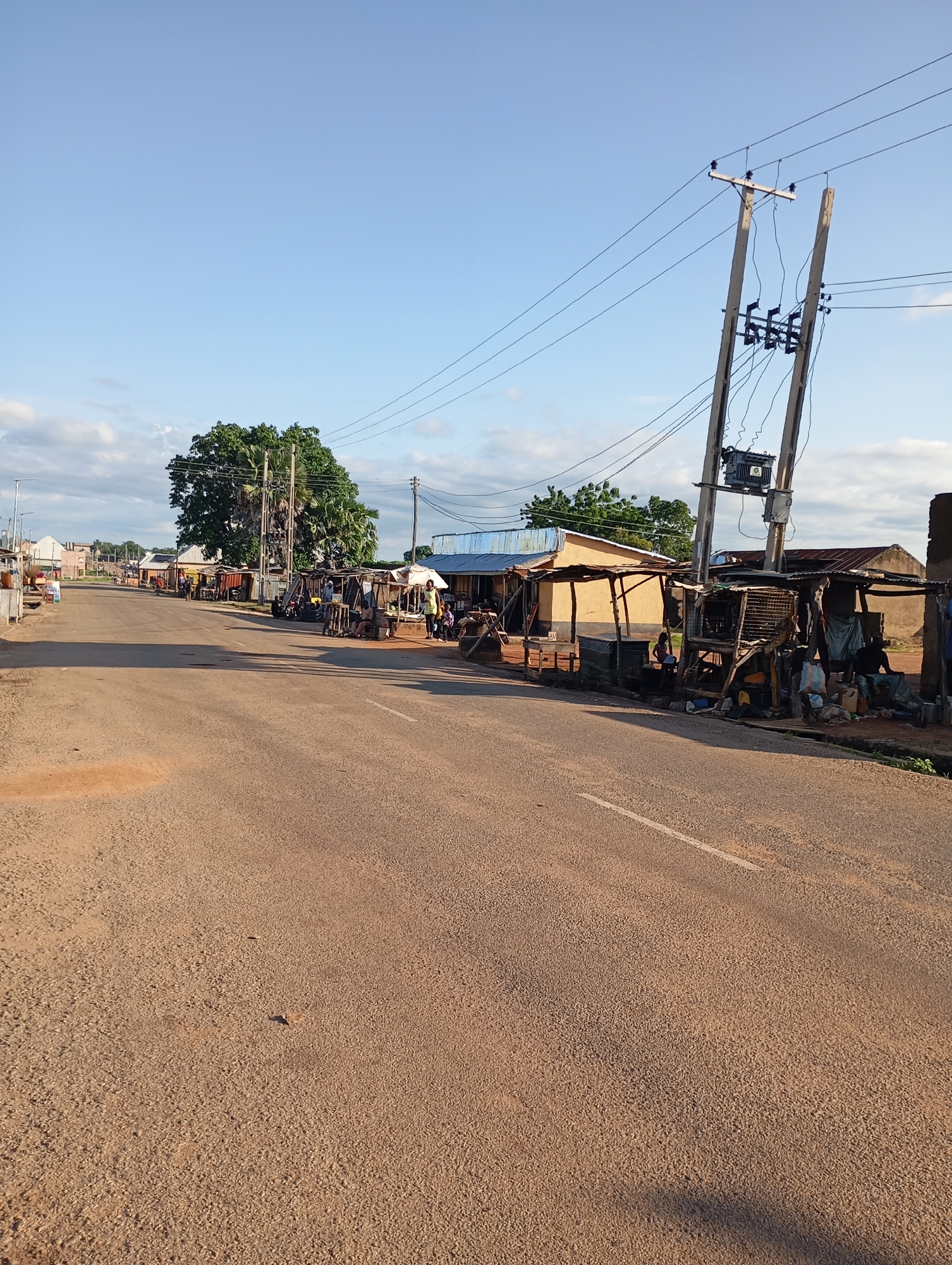 The image depicts a street scene in a rural or developing area. The road is paved and appears to be a main thoroughfare. On the right side of the road, there are several open-air market stalls and small structures constructed from wood and corrugated metal. These stalls appear to be selling goods, with people visible in and around them. One stall has a collection of bags and items piled up. A transformer is mounted on a utility pole next to the stalls, with power lines stretching across the scene.
Further down the road, on both sides, there are more buildings, some appearing to be shops or residences. A large, leafy tree is prominent on the left side of the road. Several people are visible walking along the road or in the vicinity of the stalls. The sky is blue with some scattered clouds, suggesting it is daytime. The overall impression is of a bustling community center or marketplace.