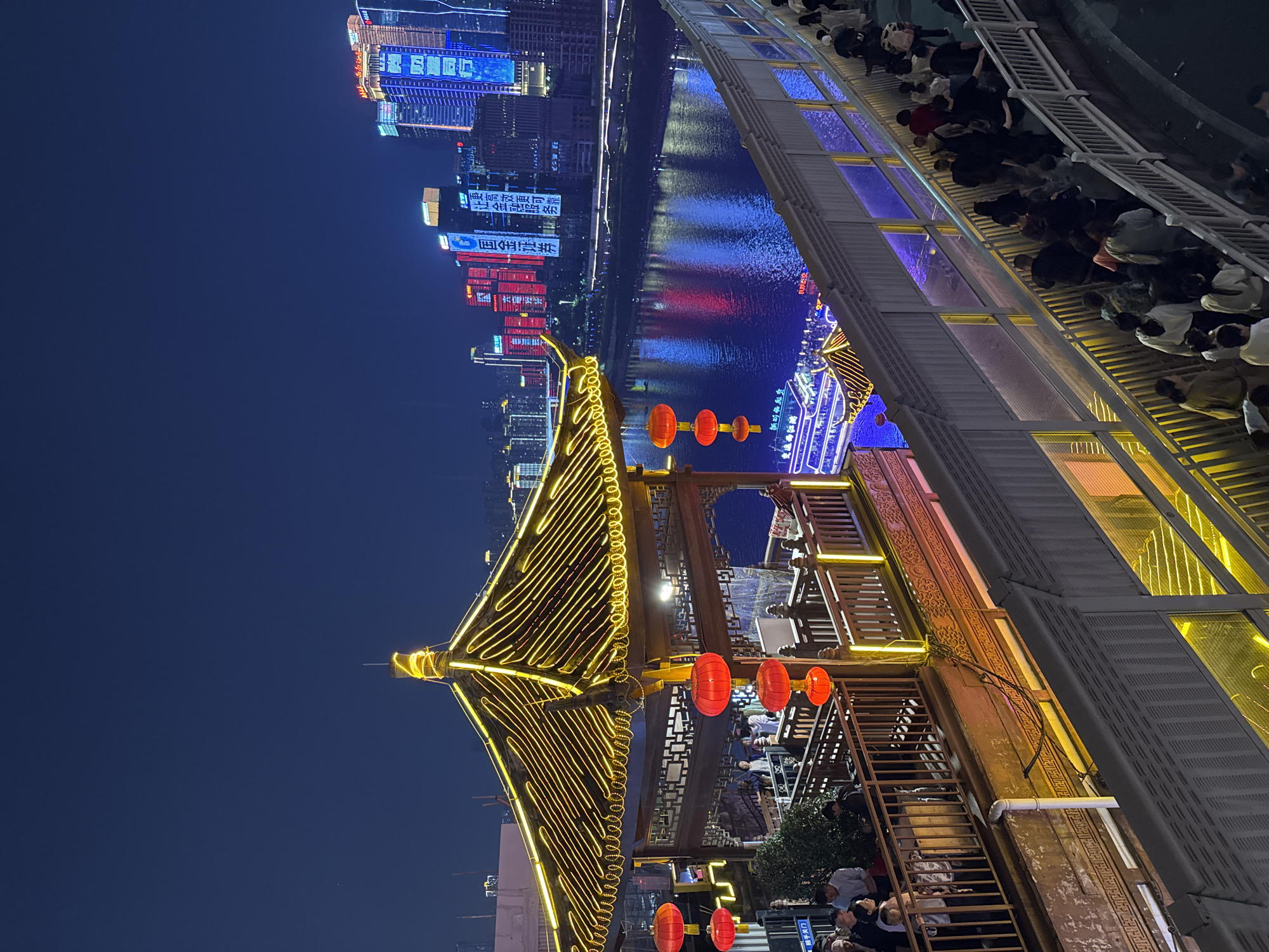 At night, a view captures the illuminated architecture and urban landscape of Yuzhong District, Chongqing, China. A traditional Chinese structure, adorned with glowing yellow lights and red lanterns, dominates the foreground. Behind it, a river reflects the lights of modern high-rise buildings. A series of people stand on a walkway.