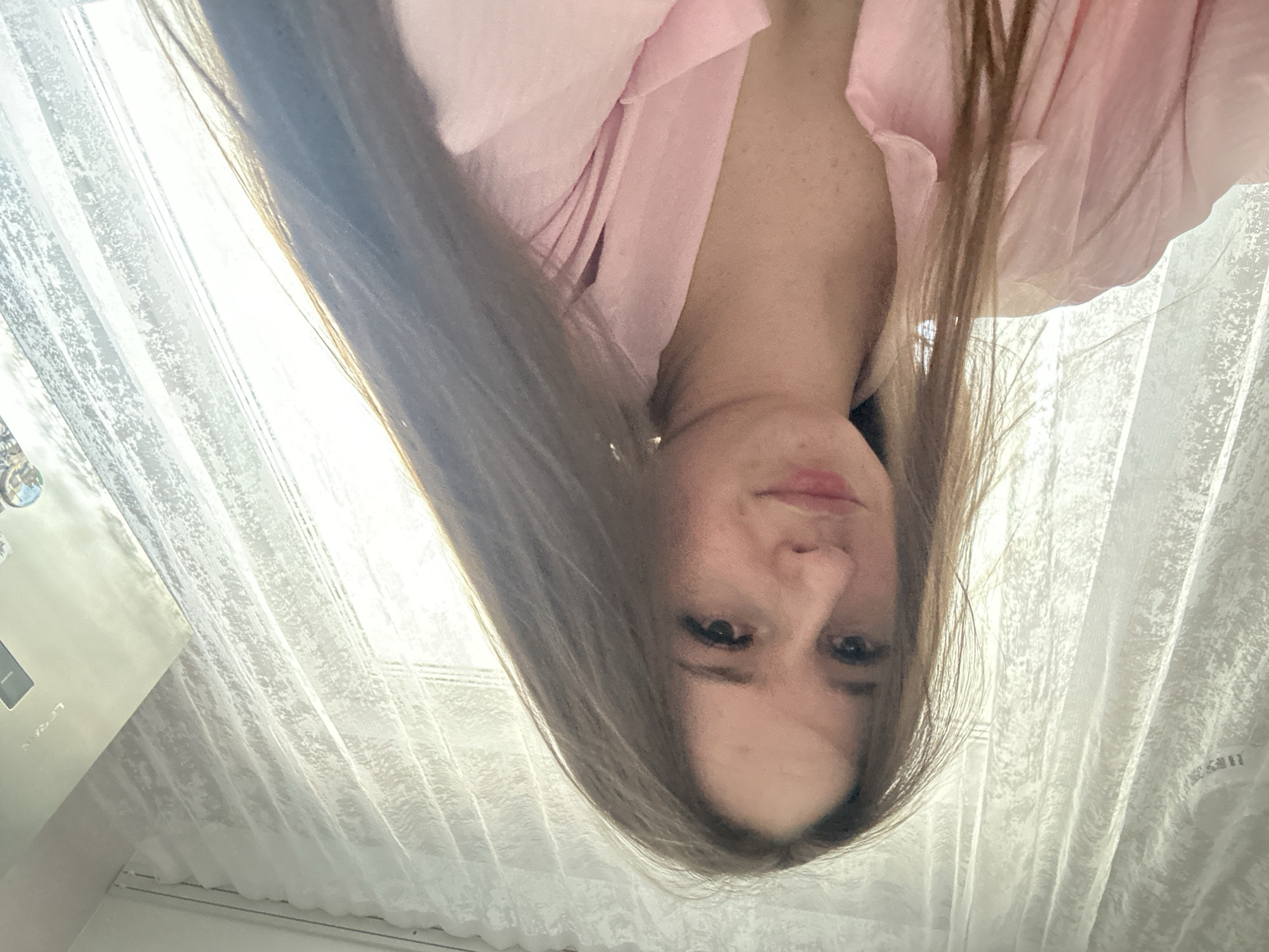 A medium-close-up image shows a woman with long, straight, light brown hair, wearing a light pink collared shirt or blouse. The photo is taken from a low angle, looking upwards at her face, which has a neutral and relaxed expression. Her eyes are open, looking directly towards the camera.
The background consists primarily of a window covered with sheer, white, textured curtains, through which bright, diffused sunlight is streaming, indicating daytime. On the far left of the frame, partially obscured by the curtains and the woman's hair, is a white electronic appliance, possibly an air conditioner unit, with the brand name "LENA" clearly visible on its surface. Faint, indecipherable text, resembling Chinese characters, can also be seen printed on the white curtain fabric in the upper right background. The overall scene conveys a casual, well-lit indoor setting.