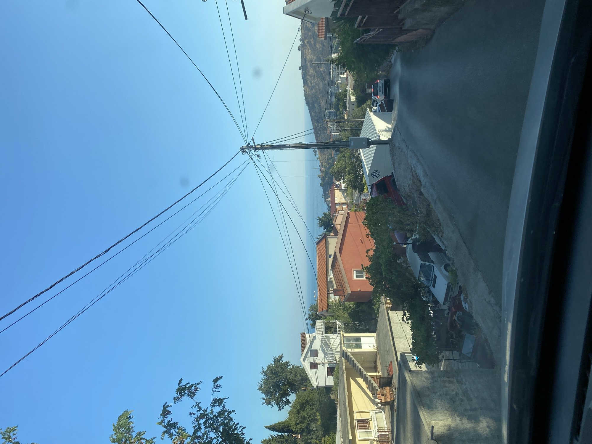 The image shows a view of a street in Sutomore, Montenegro, looking uphill towards the sea. The street is paved and curves to the right. Several houses are visible on either side of the street, with red-tiled roofs and white or colored walls. There are trees and vegetation interspersed among the houses. A utility pole with numerous wires is prominent in the middle of the frame. A few cars are parked or driving on the street, including a white SUV and a red van. In the background, the sea is visible beyond a hilly landscape with scattered buildings. The sky is clear and blue.