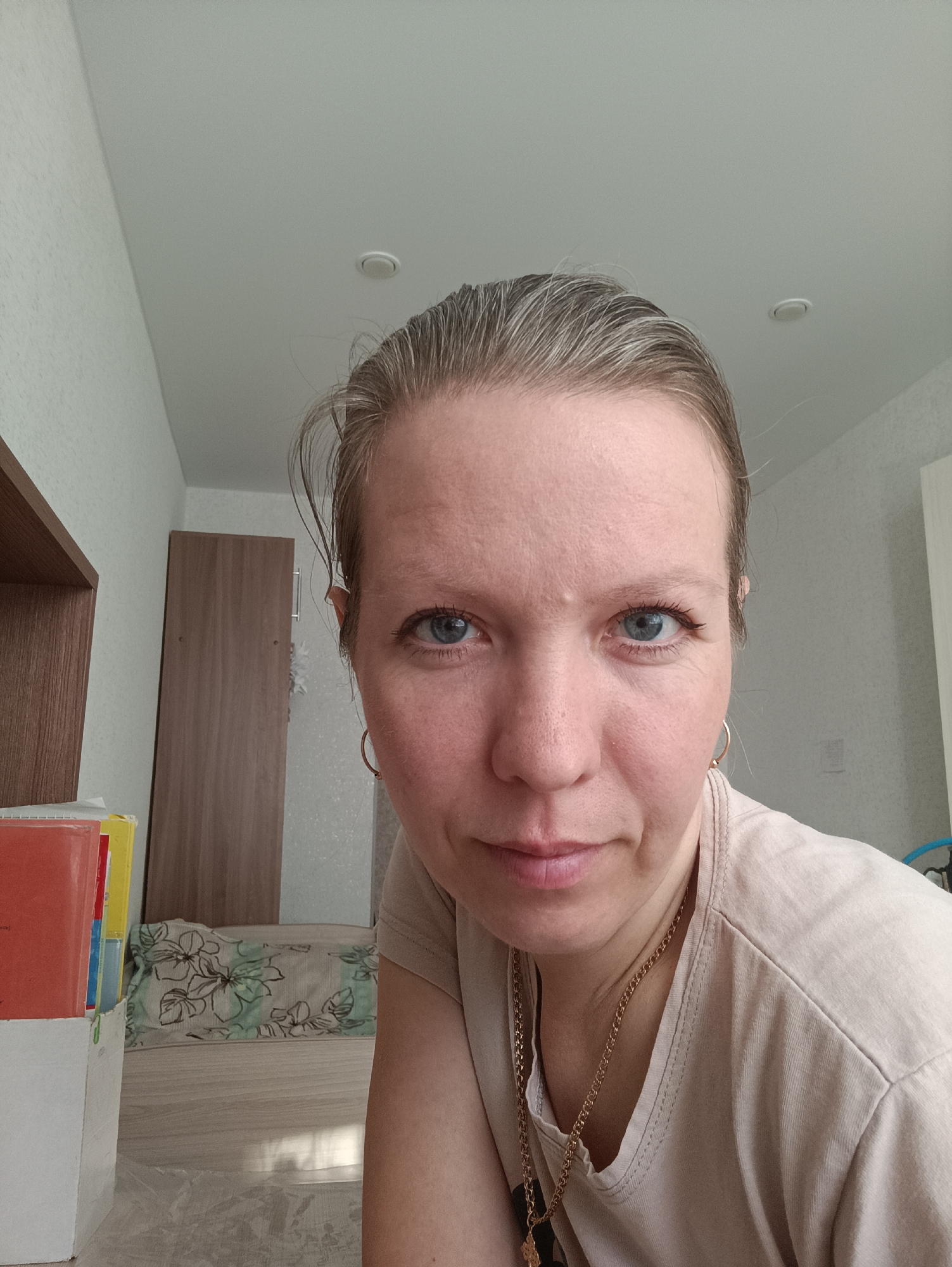 A close-up, indoor selfie depicts a woman with light complexion, blue eyes, and greying blonde hair pulled back. She is wearing a light beige t-shirt and a gold necklace with a pendant. Her face shows some wrinkles and slight redness. She is wearing gold hoop earrings. The background shows a corner of a room with light-colored walls. A stack of books is visible in a white container to the left of the frame. Behind it is a cabinet with a wooden door and a small floral patterned cushion on the floor. The ceiling is white with a round light fixture. While it is unknown if this image was taken in Chaikovsky, Russia, or anywhere else, the location cannot be determined from this image alone.