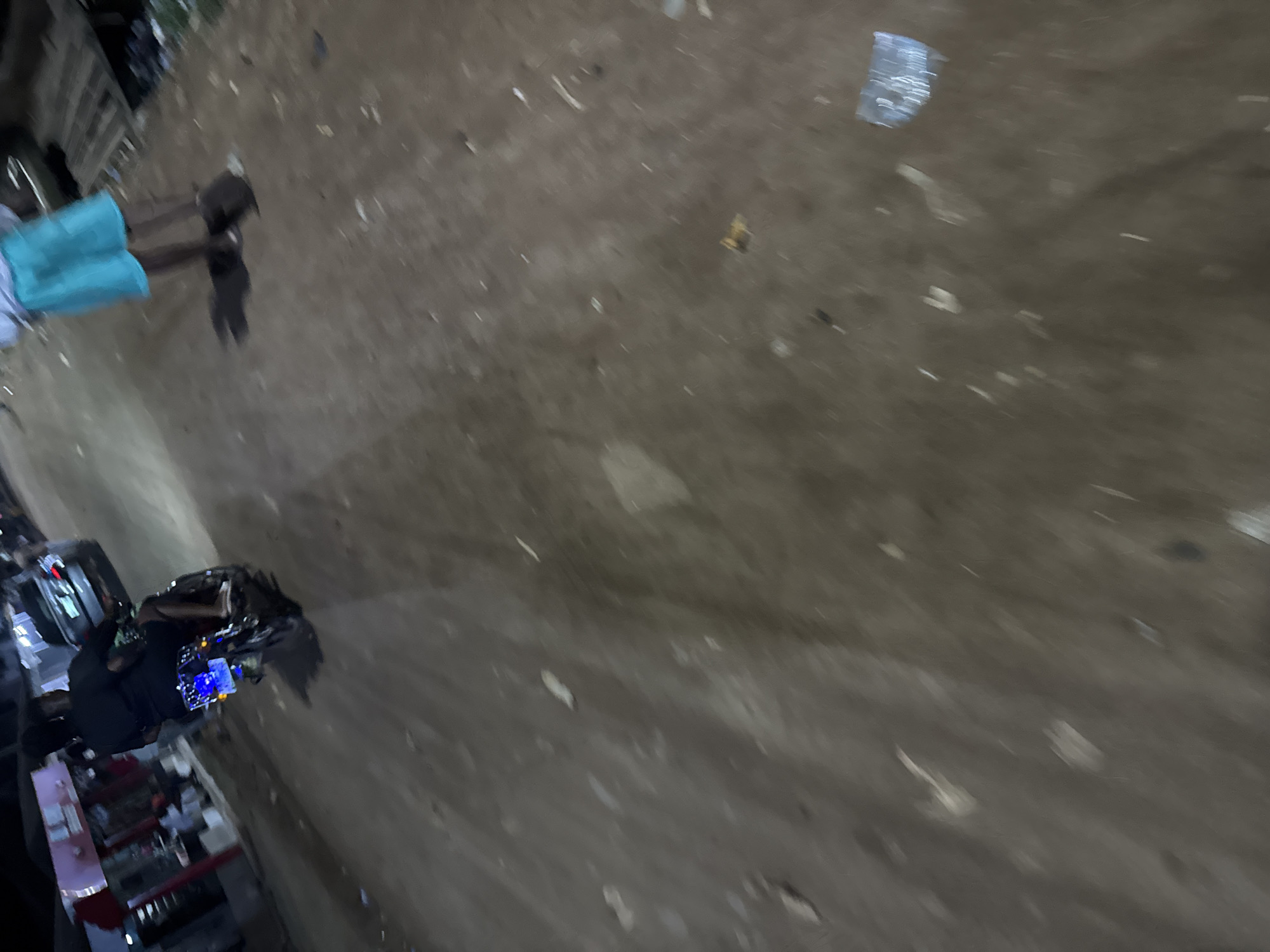 The image shows an outdoor scene in Ihiagwa, Nigeria. A person wearing a blue garment is walking past a parked motorcycle with a blue display. Debris and litter are scattered on the ground, and there are some building structures in the background. The ground appears to be made of dirt or unpaved material.