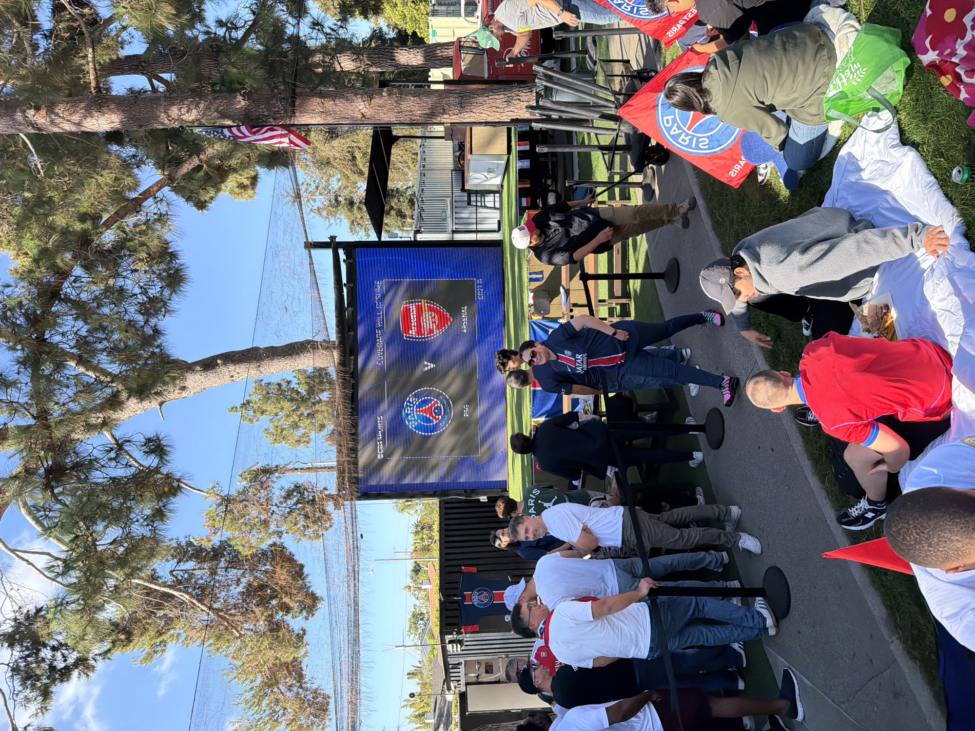 The image shows a public viewing event for a soccer game between Arsenal and PSG, likely in Los Angeles, United States. People are gathered outdoors, some standing in a line held back by stanchions, while others are seated on blankets on the grass. A large screen displays the logos of the two teams. Various individuals are visible, including adults and children, wearing different attire. An American flag is visible near a tree. The background features buildings and trees, indicating an urban or suburban setting.