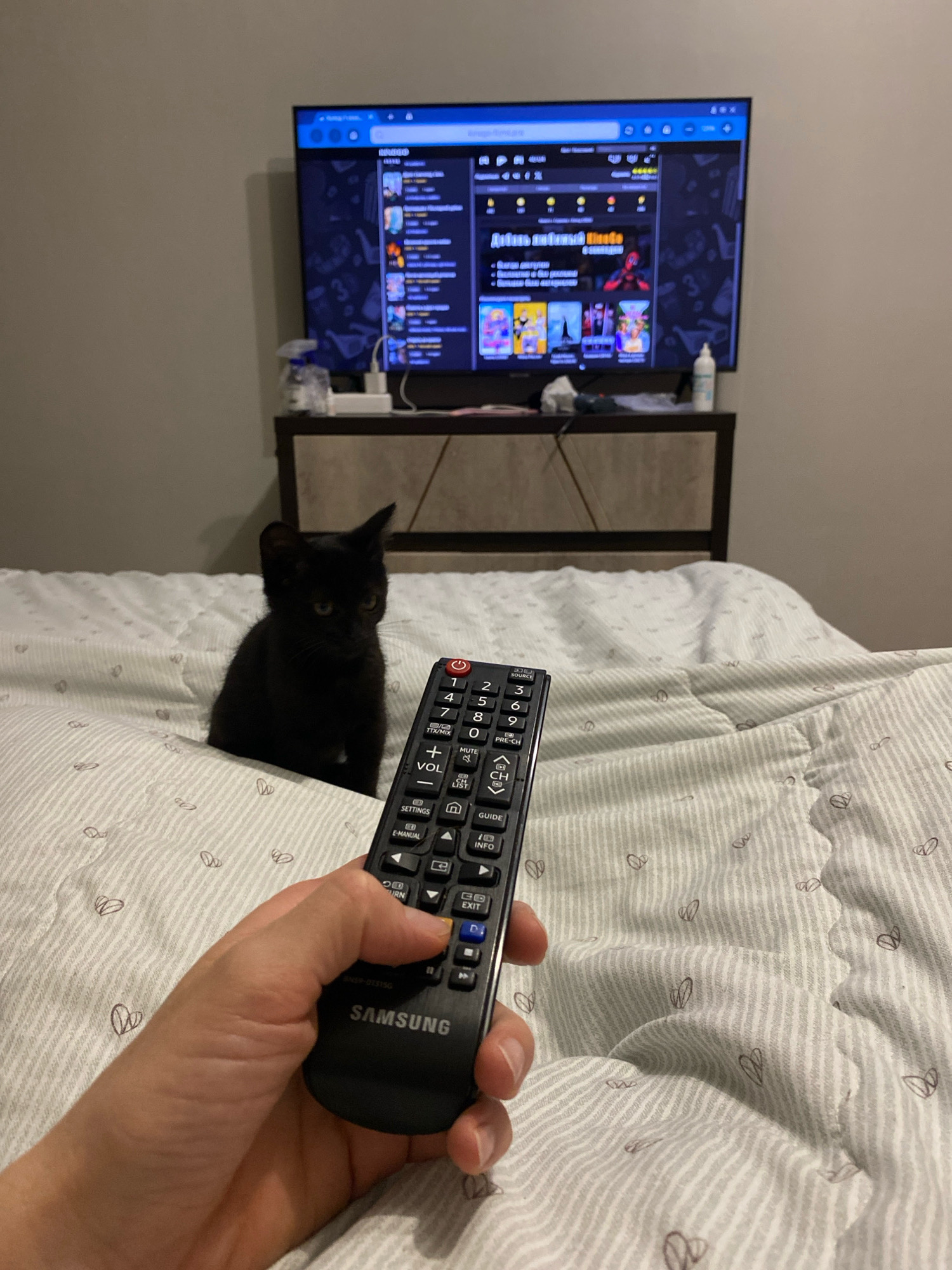 In a bedroom, on a bed with a white and gray striped comforter adorned with small heart drawings, a black kitten sits facing the viewer. In the foreground, a hand holds a black Samsung television remote, with a thumb pressing a blue button. The remote has various buttons, including numerical keys, volume controls, and a channel up/down button. Behind the kitten, a dark wooden dresser is visible, and on top of it, a television displays a colorful interface with various icons and text in what appears to be Cyrillic. A bottle of spray and a white object are placed on the dresser. The overall scene suggests a relaxed, domestic setting. The city of Zhlobin, Belarus, is not depicted in this image.