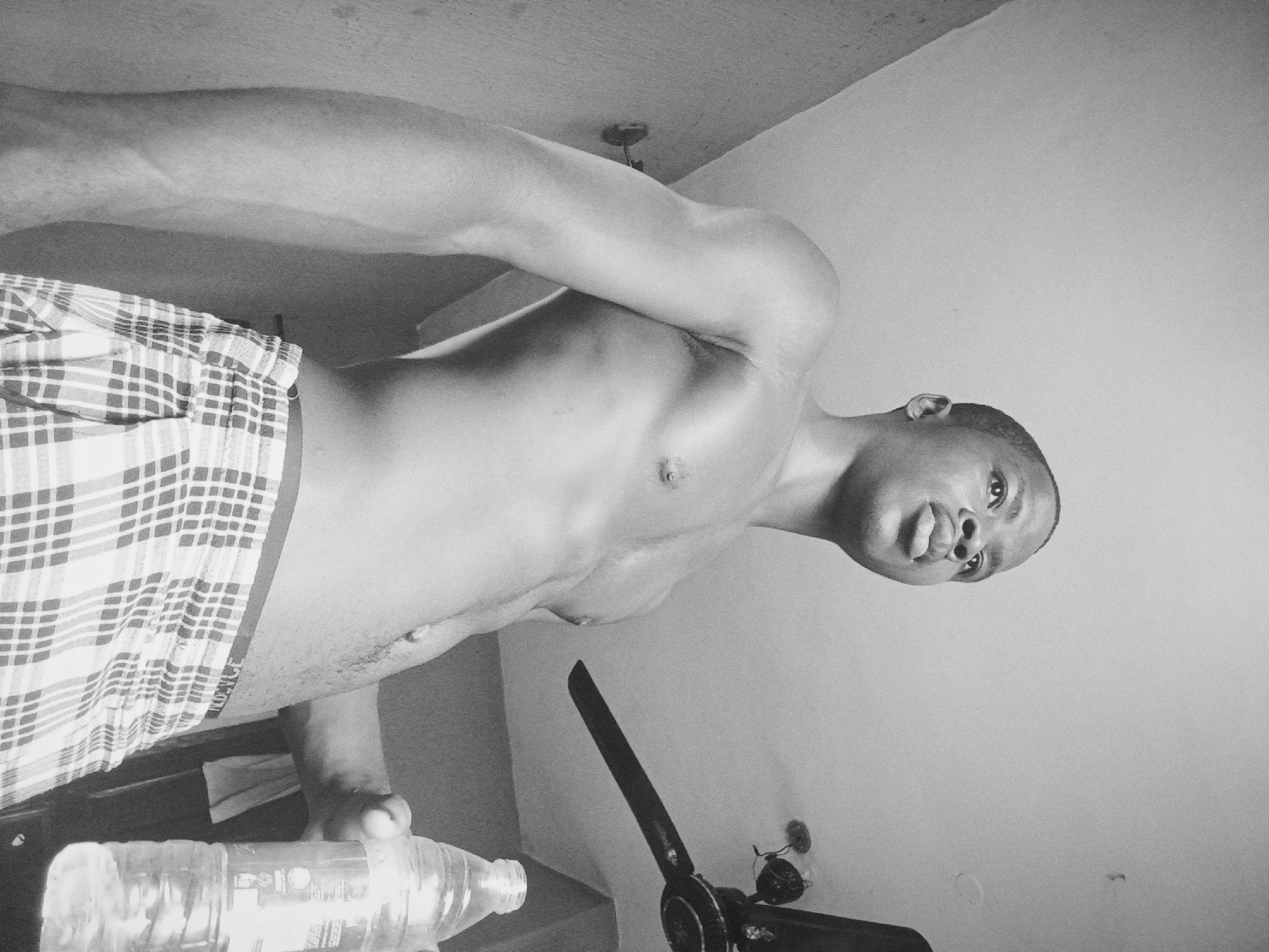 The image is a black and white, low-angle shot of a shirtless man wearing plaid shorts. He is holding a water bottle in his left hand. A ceiling fan is visible in the lower right corner, and a ceiling light is in the upper center of the frame. The background appears to be a plain wall. Based on the provided information, the location is in Nkpor, Nigeria, although there are no visual cues to verify the exact location.
