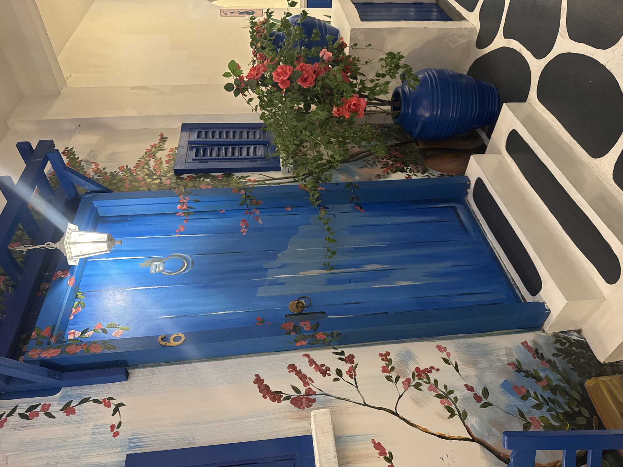 The image depicts an indoor mural reminiscent of Greek island architecture, possibly located in Denpasar, Indonesia, given the prompt's context. A vibrant blue, stylized door dominates the scene, adorned with painted flowers climbing along its frame and surface. A blue window frame is positioned above the door.
Two large, blue pots overflowing with leafy greenery and red roses are strategically placed above the door and to the right, adding depth and color. The background features white walls with stylized black and white accents.
A hanging light fixture is visible near the upper left corner of the door. The overall composition creates a faux outdoor scene, likely intended as a decorative element within an indoor space.