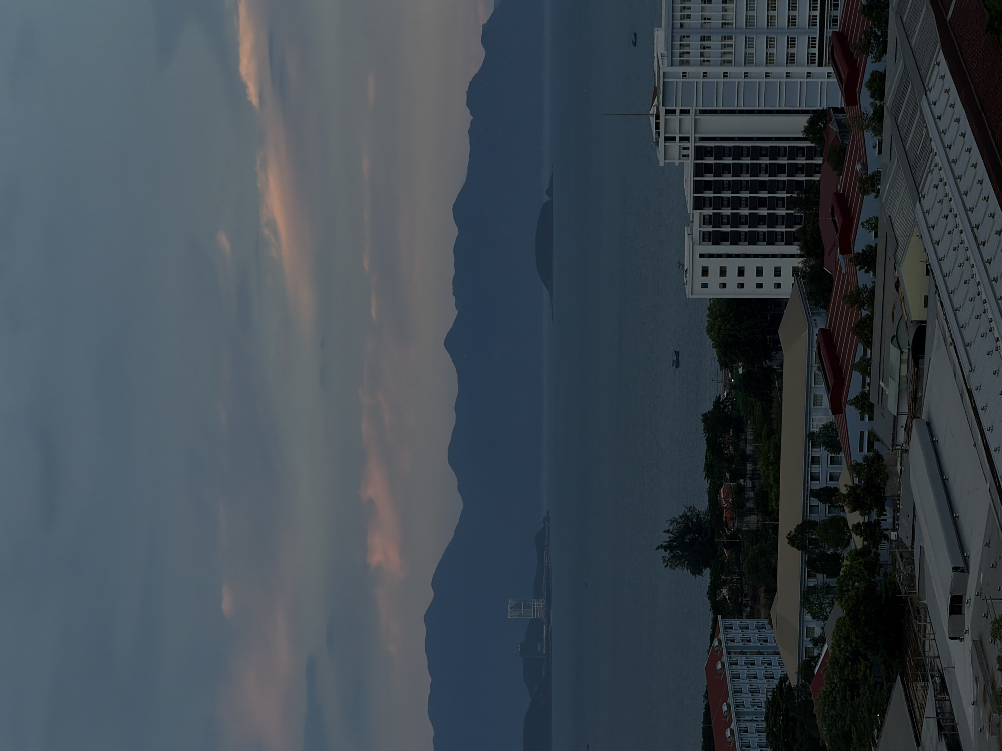 This image depicts a coastal city scene in Nha Trang Ward, Vietnam, likely during either dawn or dusk due to the soft, diffused lighting and the muted colors of the sky. The orientation of the image is vertical.
The main subjects are a cityscape and a natural landscape. In the foreground, several buildings are visible, featuring modern architecture with white and grey facades, interspersed with red-tiled roofs and lush green trees. One building stands out with its numerous windows and balconies. Further into the scene, a body of water, likely the sea, stretches out to meet a range of dark, silhouetted mountains in the background. The water appears calm, with a few small boats dotting its surface. A distinct, low-lying structure or island with a building on it is also visible in the water, positioned between the city and the mountains.
The sky is a prominent element, filled with soft, wispy clouds that are tinged with hues of pink and orange, indicative of the twilight hours. The overall mood is serene and tranquil, reflecting the peacefulness of the coastal environment at this time of day. The weather appears to be clear and calm.
There is no visible text in the image. The scene captures the harmonious coexistence of urban development and natural beauty.