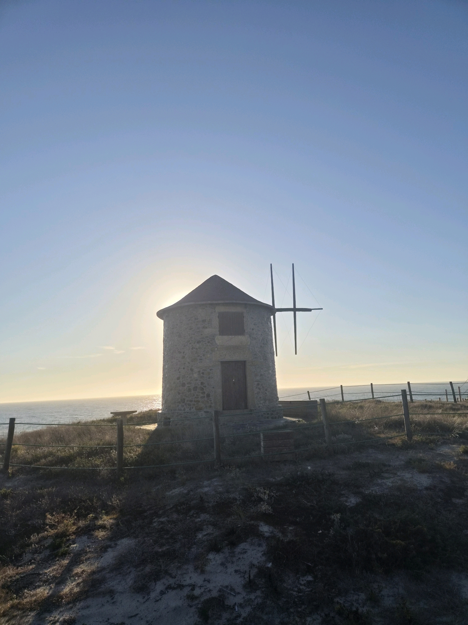 The image shows a stone windmill with a conical roof, located on a grassy knoll overlooking the ocean. The sun is setting or rising behind the windmill, casting a warm glow on the scene. A wooden fence surrounds the windmill, and a bench is visible to the left. The sky is clear and blue. This windmill is one of the windmills of Esposende, Portugal, which are a series of traditional windmills located along the coast of Esposende. They are a popular tourist attraction and a symbol of the town's maritime heritage.