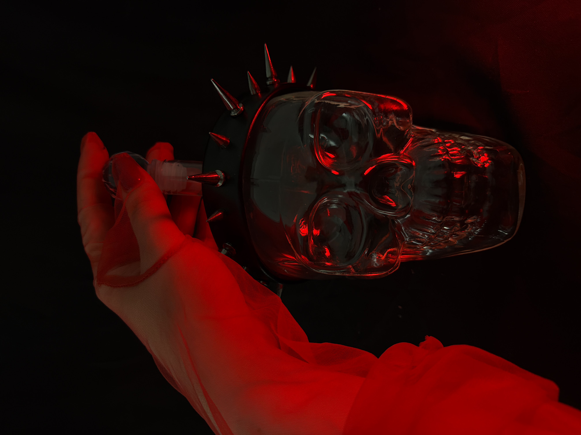 A human hand, clad in a sheer, red-tinted glove, is seen from the side, reaching towards a clear glass skull-shaped bottle. The bottle is adorned with a black spiked collar around its upper portion, with several sharp, conical spikes pointing outwards. The skull's eye sockets and facial features are clearly visible, reflecting the red light. The background is entirely black, creating a stark contrast and emphasizing the illuminated objects. The scene appears to be a still life, possibly set up for a photograph, with no indication of a specific urban location. The hand is in the process of holding or adjusting the bottle.