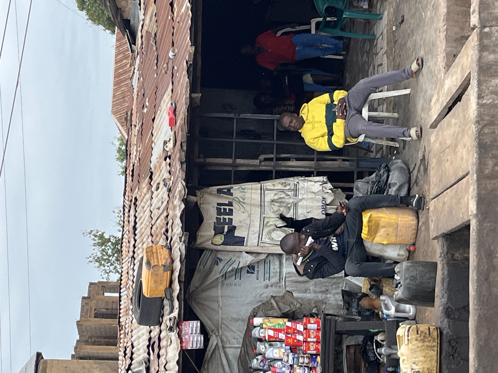In Gura Top, Nigeria, a street scene unfolds under an overcast sky. A makeshift shop, constructed with corrugated metal roofing, stands prominently. On the roof, a deflated tire and a weathered yellow plastic container are visible. In front of the shop, two men are seated. The man on the left, wearing a black jacket and blue jeans, is on the phone. The man on the right, in a yellow and green hooded sweatshirt and gray pants, sits with his hands clasped. Behind them, the shop interior is dimly lit, with several people visible. To the left of the shop, a display of various liquid products in bottles and cans is arranged on a wooden table. Several large plastic containers, some yellow and some dark-colored, are placed on the ground in front of the shop. A partially constructed building with concrete blocks is visible in the background, suggesting ongoing development in the area. The ground is a mix of dirt and concrete, with some debris scattered around.