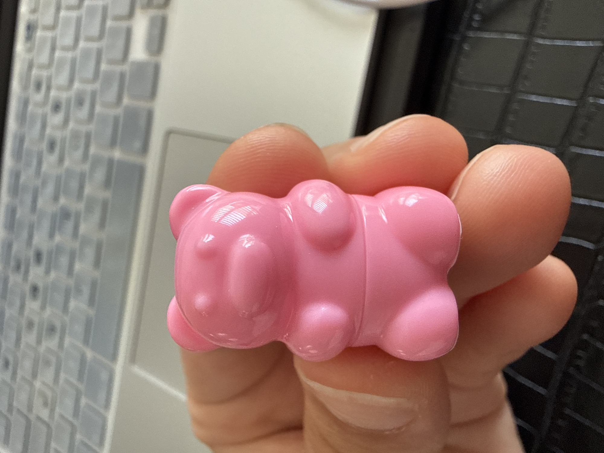 A close-up shot shows a person's hand holding a small, pink, bear-shaped object. The bear is shiny and appears to be made of plastic or a similar material. The background is blurred, but it appears to be a keyboard on a laptop. The lighting suggests an indoor setting. There is no information in the image to indicate a location in Marum, Netherlands.