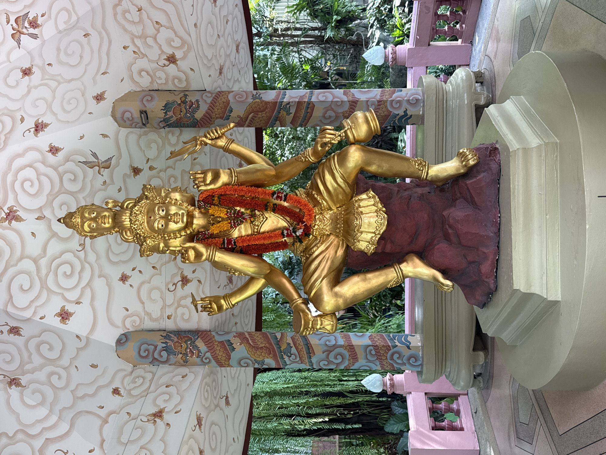 A golden statue of a multi-armed deity is the central element of the image. The statue is adorned with a garland of orange and yellow flowers and is positioned on a maroon base. The deity has multiple faces and is holding various objects in its hands, including a knife and a pot. The statue is situated inside a structure with a ceiling decorated with cloud patterns and floral motifs. Pillars painted with dragon designs support the structure. Beyond the pillars, lush green foliage is visible. The scene appears to be located in Samrong Nuea Subdistrict Municipality, Thailand, based on visual cues associated with Thai religious iconography and architecture.