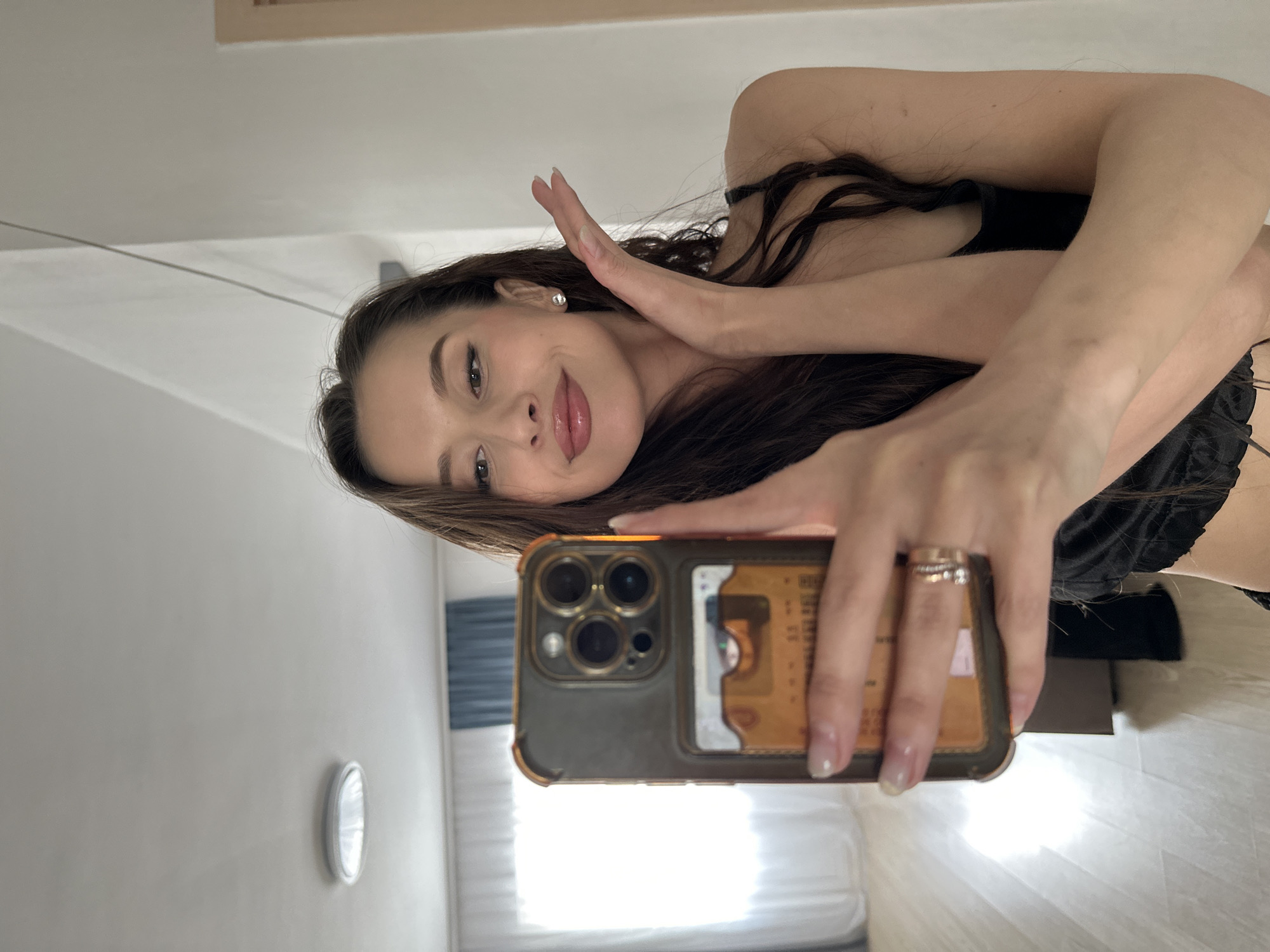 A woman is taking a selfie with her phone. She has long brown hair, light skin, and is wearing a dark top. She is holding up her left hand near her face. The phone has a dark case with visible credit cards on the back. The background shows a bedroom with a bed and a window with white curtains. The location appears to be indoors. There is no indication that the picture was taken in Yekaterinburg, Russia.