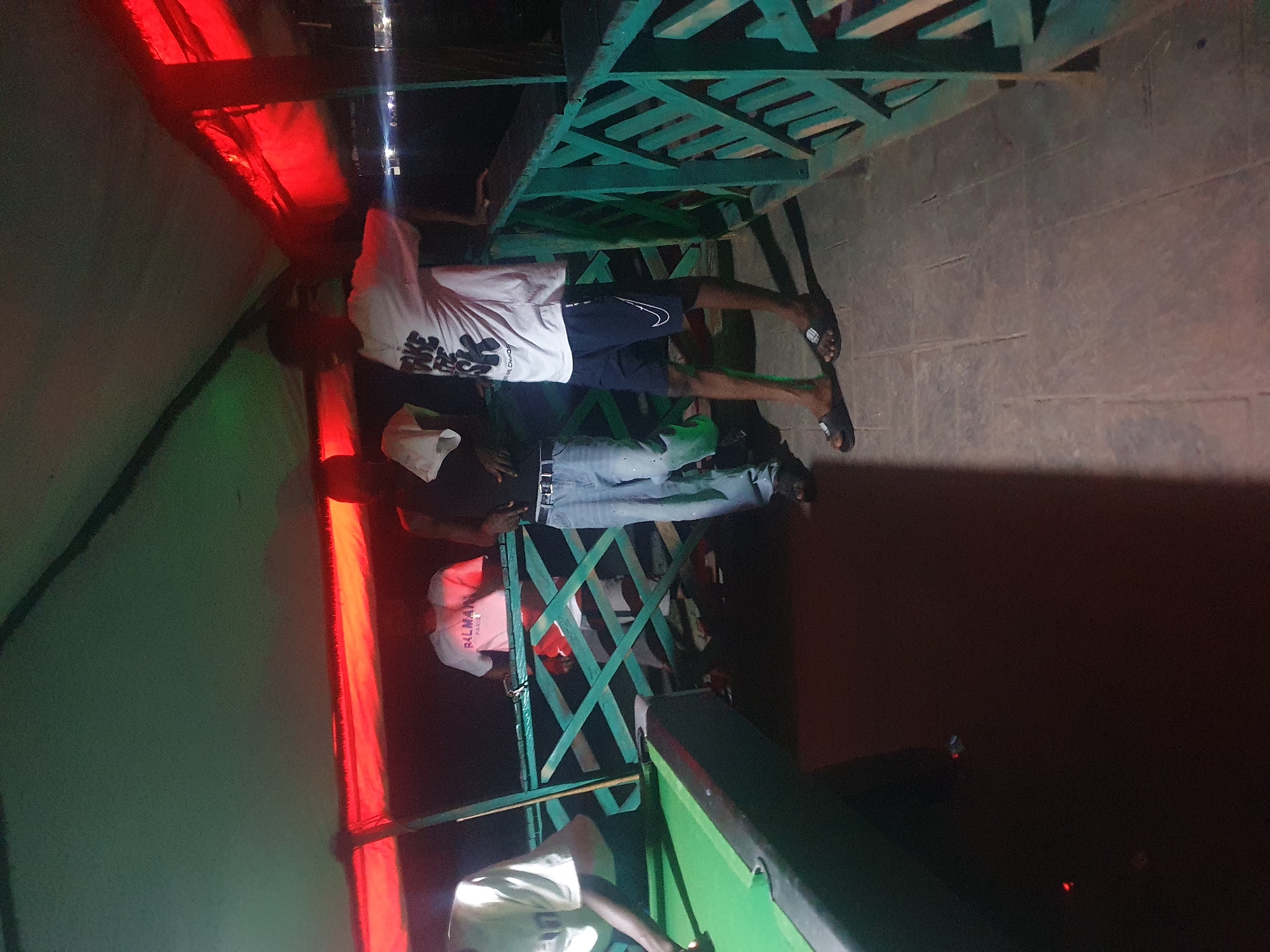 The image shows a group of people gathered around a pool table, possibly in a recreational area or club. The setting appears to be dimly lit, with red and green lighting contributing to the ambiance. Several individuals are visible, some standing and others possibly seated or leaning.
One person in the foreground, wearing a white t-shirt with black lettering, is facing away from the camera, towards the pool table. Another person, in the middle ground, is wearing a white t-shirt with "BALMAIN PARIS" printed on it in black. They are also facing towards the pool table. Other individuals are partially visible, engaged in conversation or observing the game.
The pool table itself is green and appears to be in use, with a cue stick resting on it. The surrounding area is characterized by a green, lattice-like structure, possibly part of a railing or a decorative element. The floor is paved, appearing to be made of concrete or stone.
The time of day is likely evening or night due to the artificial lighting and the dark surroundings. The overall mood seems relaxed and social, suggesting a casual gathering or a game night. The location context of Lokoja, Nigeria, is provided, but no specific landmarks are discernible within the image itself.