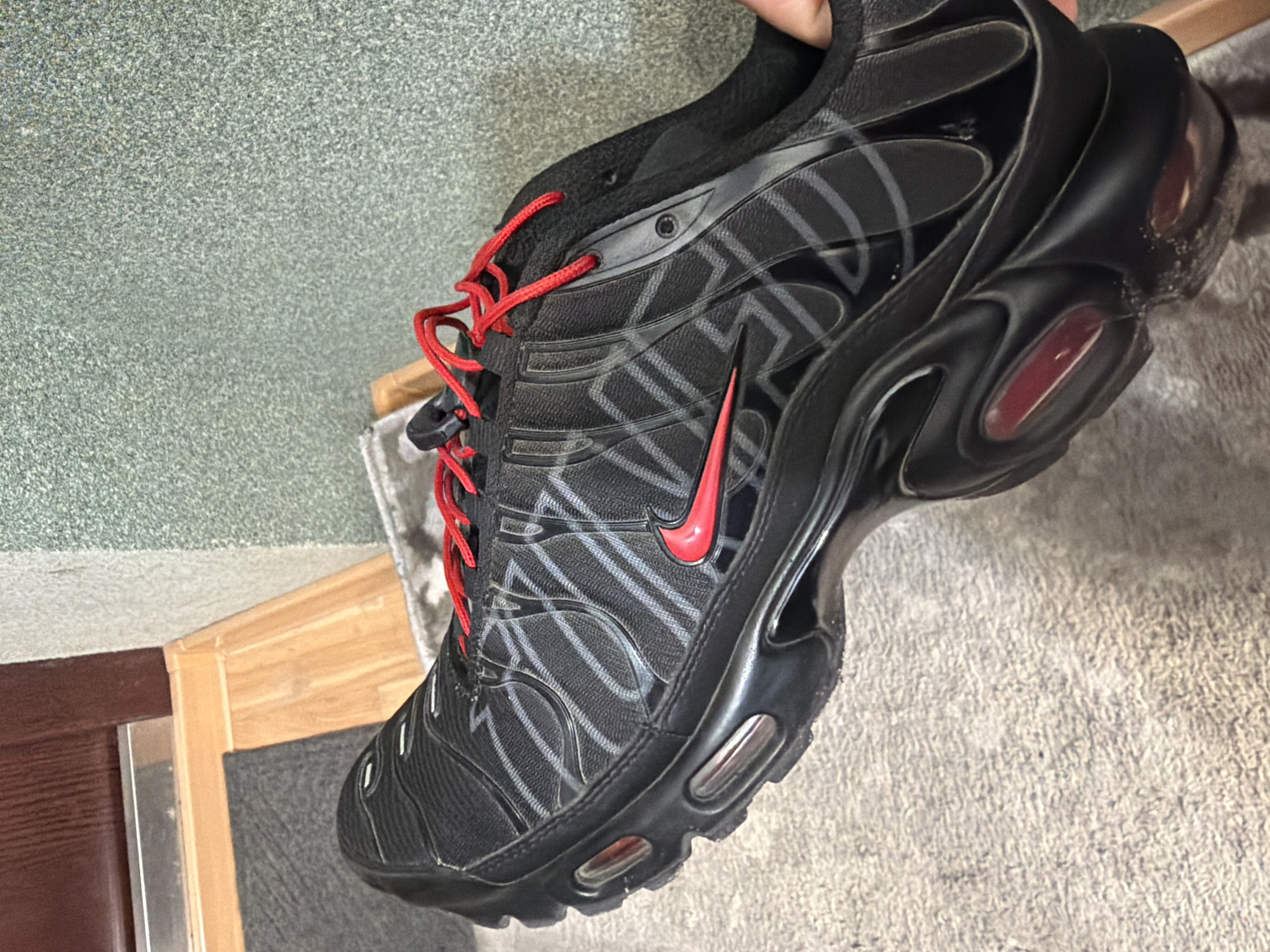 A black and grey Nike Air Max Plus TN sneaker with red laces and a red Nike swoosh logo is held up in front of a carpeted floor, a wooden door frame, and a greenish-grey wall. The location in the city of Kovel, Ukraine is not discernible from the provided image.