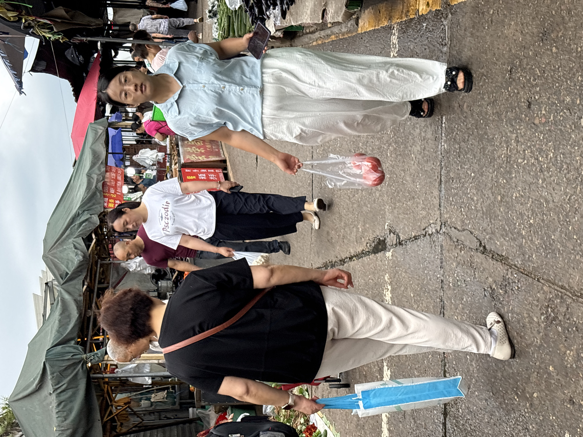 The image depicts an outdoor street scene, likely a market or shopping area in Jiangjin, China. The foreground features a person with short curly hair and a black shirt, wearing a shoulder bag, holding a blue bag, and walking on a wet street. In the middle ground, two other individuals are visible: one in a white t-shirt with the word "Pszczolin" and another one in a maroon shirt. In the background, there's a woman in a blue blouse looking at the camera, wearing glasses and holding a mobile phone in one hand and a bag of red items, likely fruits, in the other. The area is crowded with people and stalls covered with awnings, possibly selling produce or other goods. The street appears wet, indicating recent rain or cleaning. There are signs with Chinese characters on the stalls.