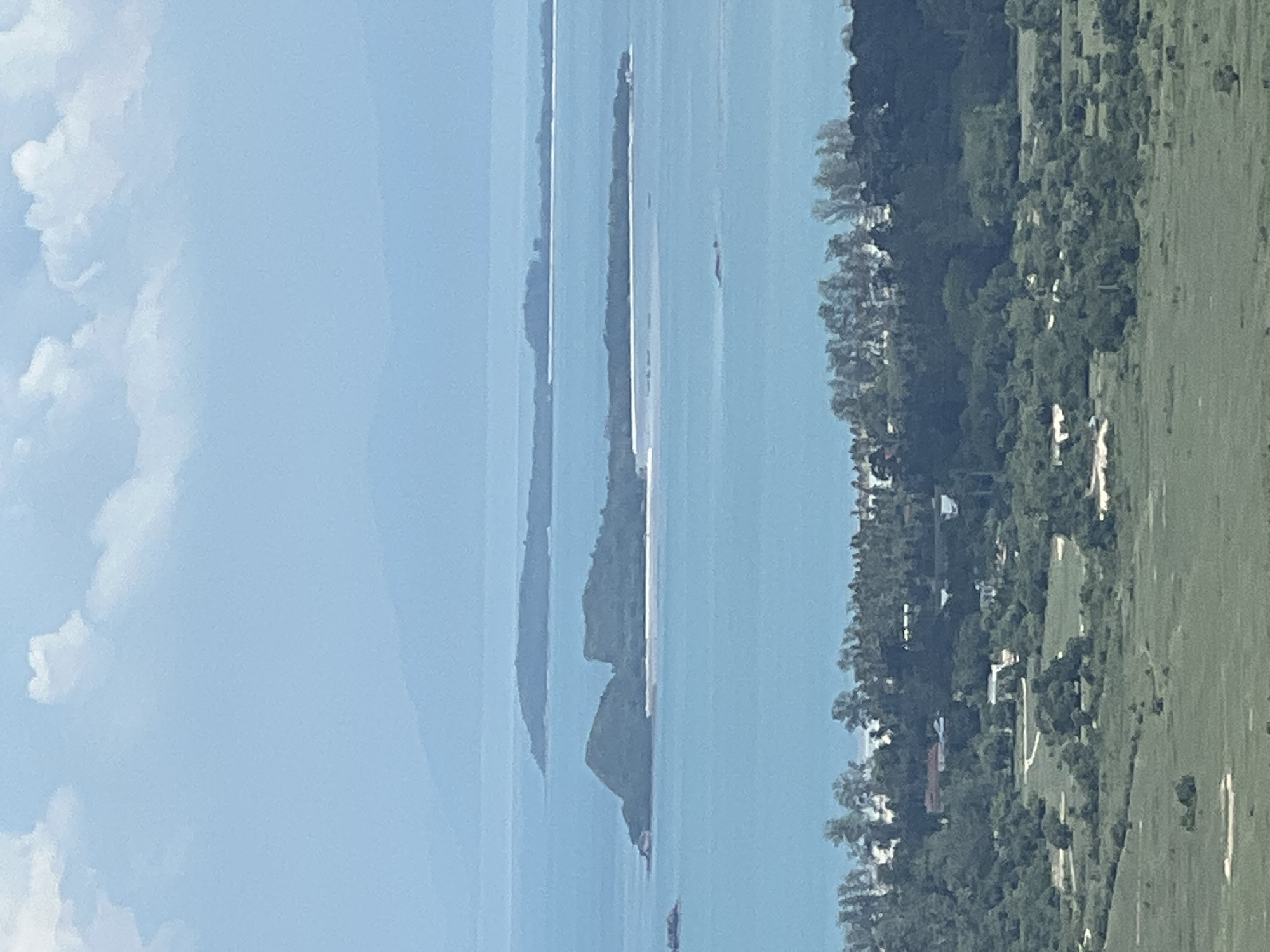 This is an aerial, wide-angle photograph taken from a high vantage point, likely a hill or mountain, overlooking a coastal landscape. The scene is dominated by the ocean, which takes up approximately two-thirds of the frame, and a distinct horizon line. In the foreground, a lush green, undulating landscape with scattered trees and some structures is visible, suggesting a rural or semi-rural area. The mid-ground features a dense line of trees along the coastline, interspersed with some buildings, indicating a populated coastal settlement. Beyond this, several islands of varying sizes and elevations are scattered across the tranquil, blue ocean. The largest island in the mid-distance has a rugged, mountainous terrain and a white sandy beach. Further in the distance, more islands and landmasses are faintly visible, fading into a hazy blue sky. The sky itself is partly cloudy, with soft, white clouds against a pale blue background. The overall impression is one of a serene tropical environment. The image is consistent with the geography and scenery found in Baan Thurian, Thailand, a location known for its coastal beauty and islands.