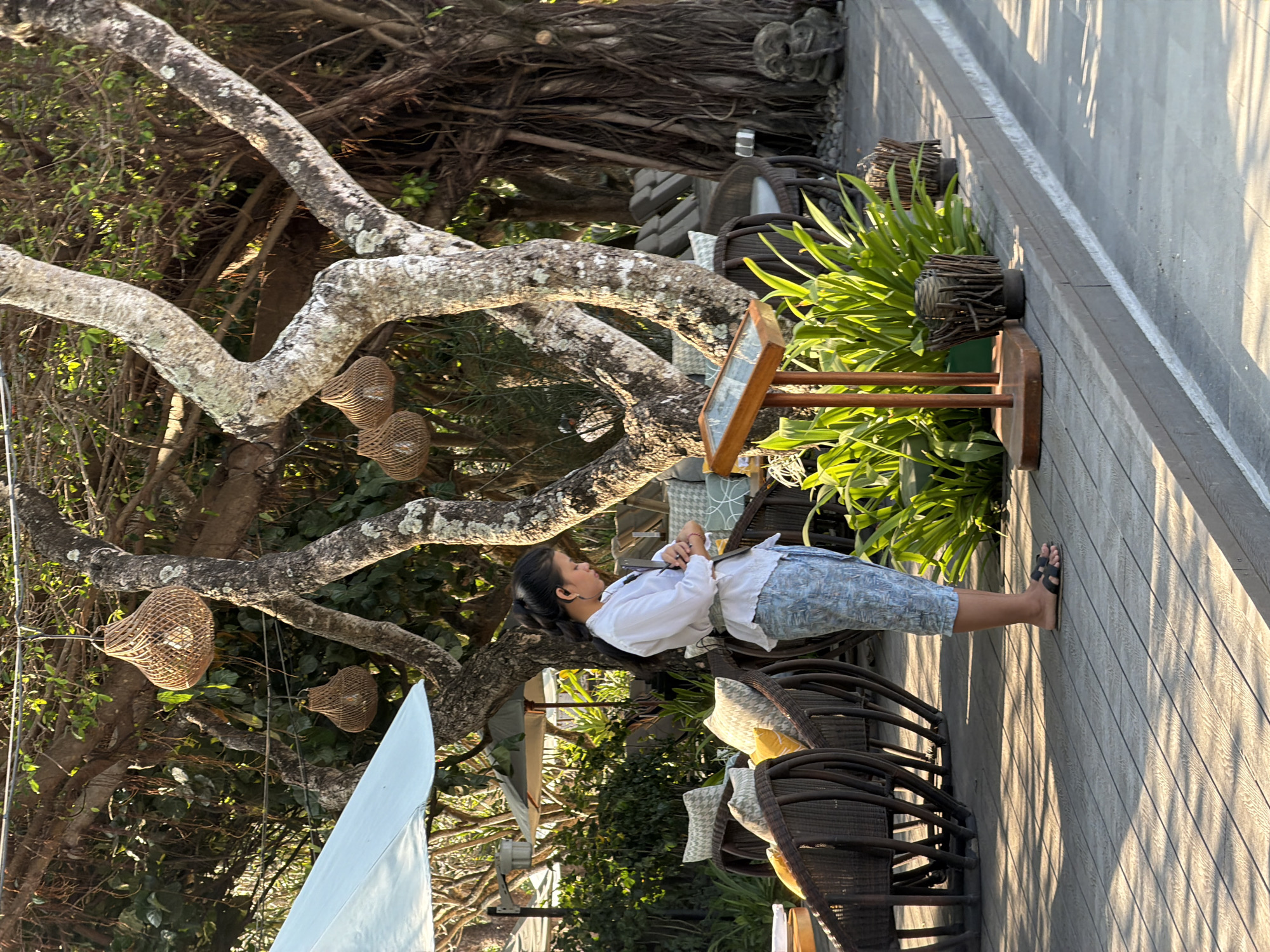The image depicts an outdoor seating area, possibly a restaurant or cafe, with a young woman standing to the left of a wooden display stand. She is wearing a white long-sleeved blouse and patterned pants, with her hair pulled back in a ponytail. She is looking down at what appears to be a small electronic device in her hands. The seating area features rattan chairs with cushions and is shaded by a large tree with hanging woven lamps. In the background, there are more chairs, umbrellas, and lush green foliage. To the right of the woman, the display stand holds a menu or information, surrounded by plants. The ground is paved with wooden planks that cast long shadows from the overhead branches and lamps, suggesting it is daytime. The scene is set in Denpasar, Indonesia, a city known for its vibrant culture and tropical climate, which is reflected in the lush vegetation and outdoor dining setup.