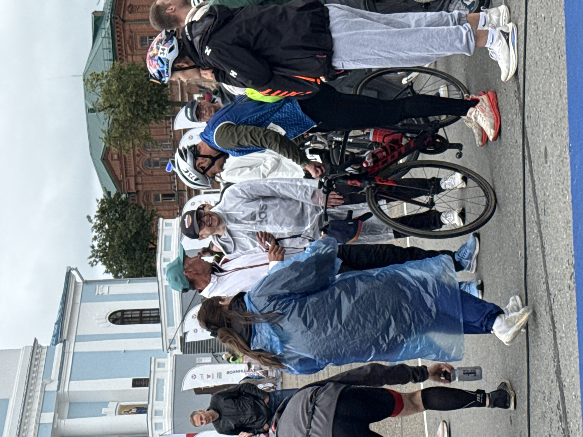 A group of people, some wearing cycling gear and helmets, are gathered around bicycles in what appears to be a public square in Votkinsk, Russia. A large, light blue and white building with a dome, possibly a church or municipal building, is visible in the background. Other buildings, including a red brick structure, are also present. The sky is overcast. Several individuals are conversing, and one person is holding a water bottle. Some are wearing rain ponchos, suggesting inclement weather. The ground is paved and marked with lines. The overall scene depicts an event or gathering related to cycling.