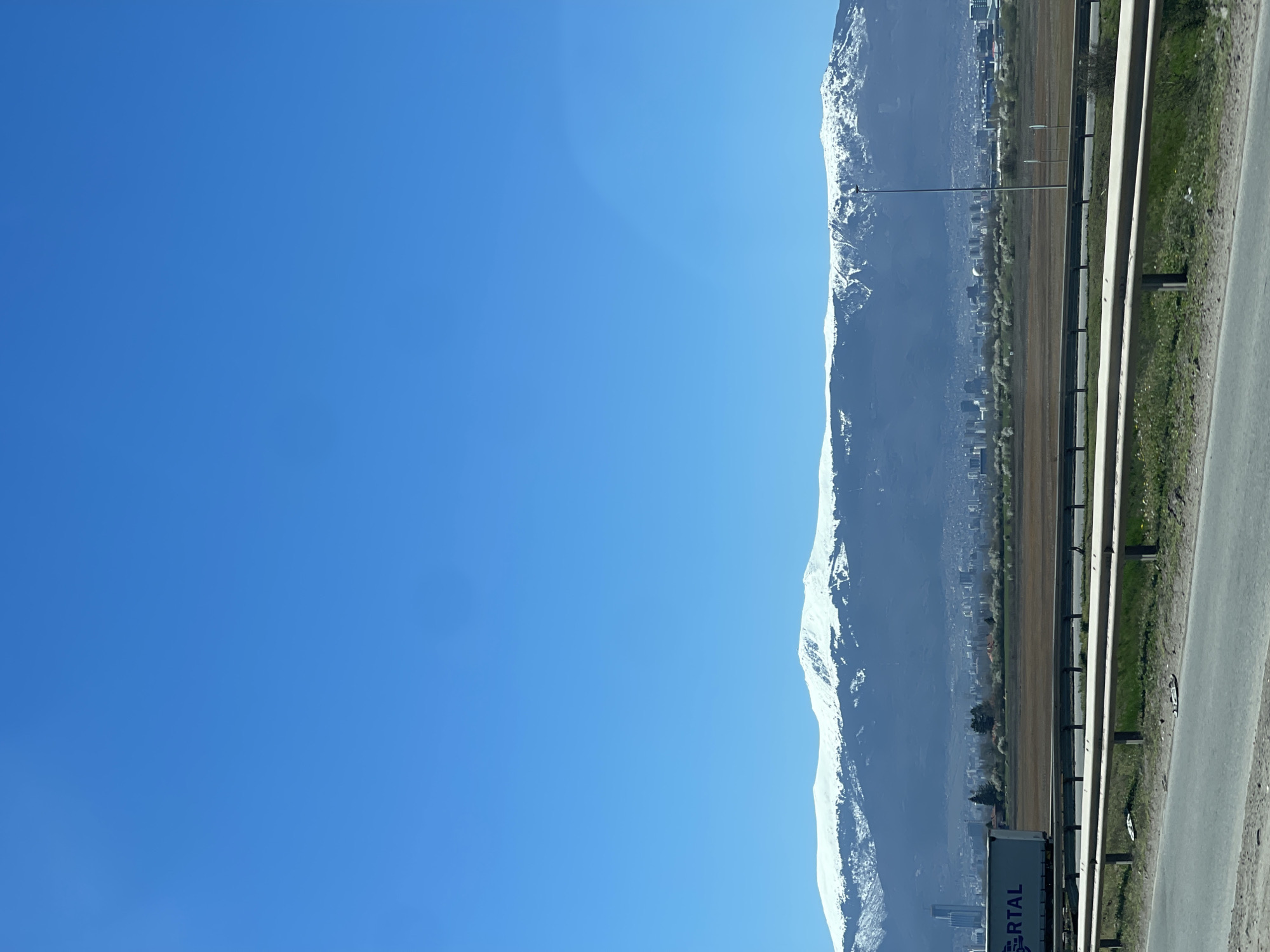 The image shows a view of the Vitosha Mountain range partially covered in snow. A highway runs along the foot of the mountain, with a city visible in the distance. Guardrails are visible along the highway. The sky is clear blue. The city is Sofia, Bulgaria.
