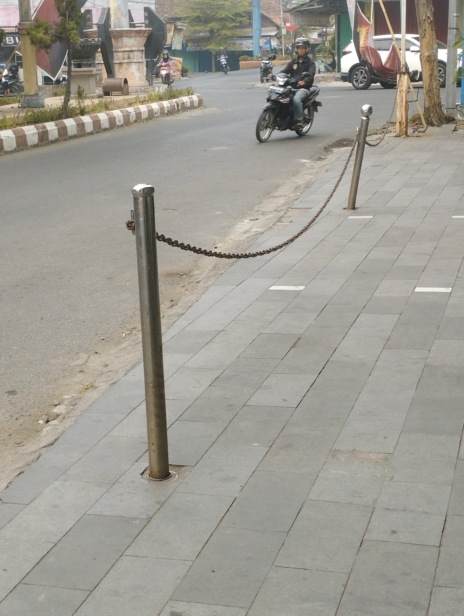 In Talang Jawa, Indonesia, a street scene unfolds with a motorcycle rider proceeding down a paved road. The rider, wearing a dark shirt, jeans, and a helmet, is on a black motorcycle. To the right of the road, a sidewalk is bordered by a metal chain fence connected to posts. The sidewalk is paved with grey tiles, and white rectangular markers are embedded in the tiles at intervals. Beyond the sidewalk, trees are visible, and buildings line the street. In the background, another motorcycle is parked near a white vehicle. The environment appears to be a typical urban street.