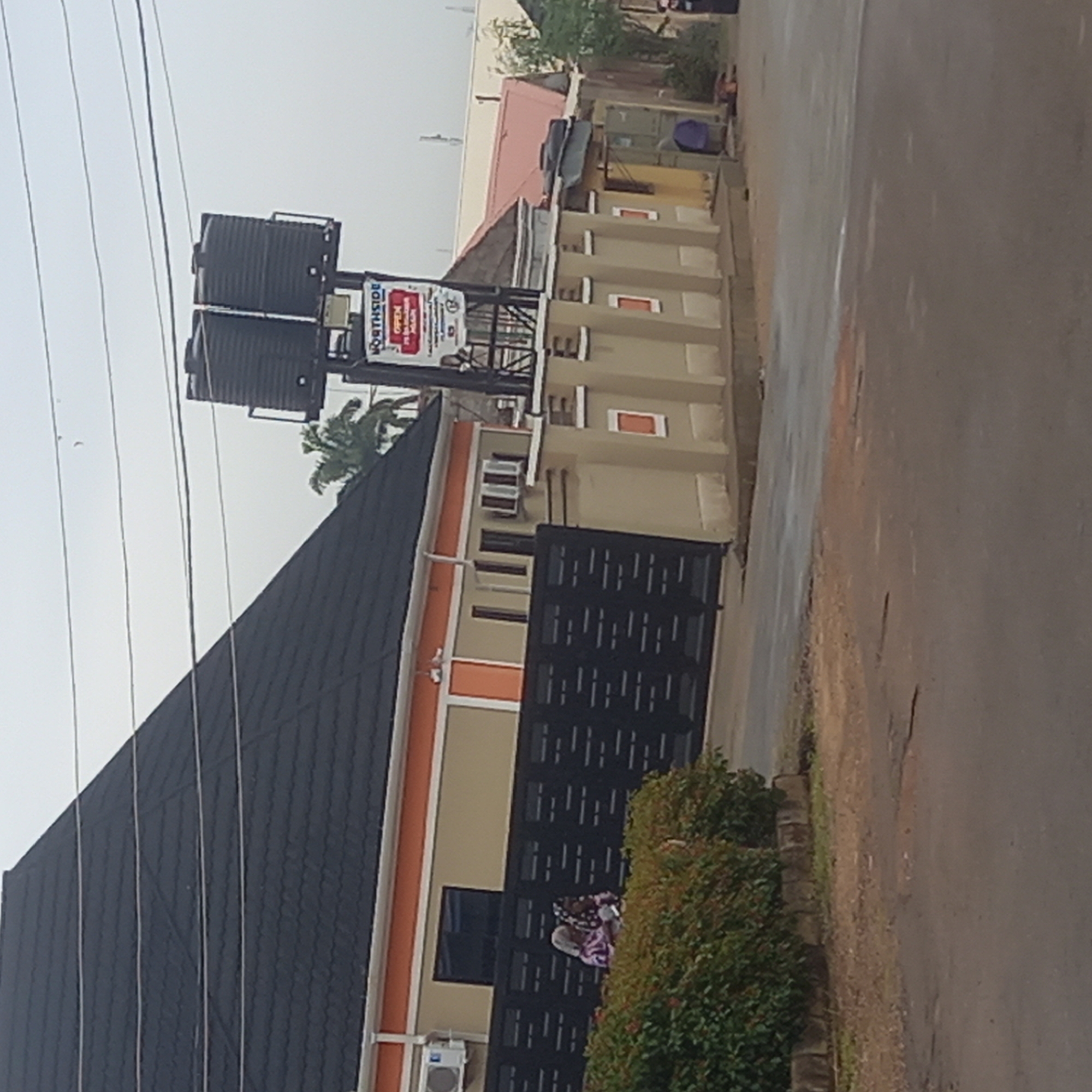 A daytime outdoor shot depicting a street scene in Barden Yana, Nigeria. Visible is a building with a dark roof and a black metal gate. The building has a beige exterior with orange accents. Attached to the building are two black water tanks mounted on a metal structure with a sign that reads "NORTHSIDE." An air conditioning unit is visible near the gate. A bush lines the building along the street. The street is paved and shows signs of wear. Over the building is a mix of power lines and a palm tree. Other buildings are visible in the background. An indiscernible object is lying on the gate.