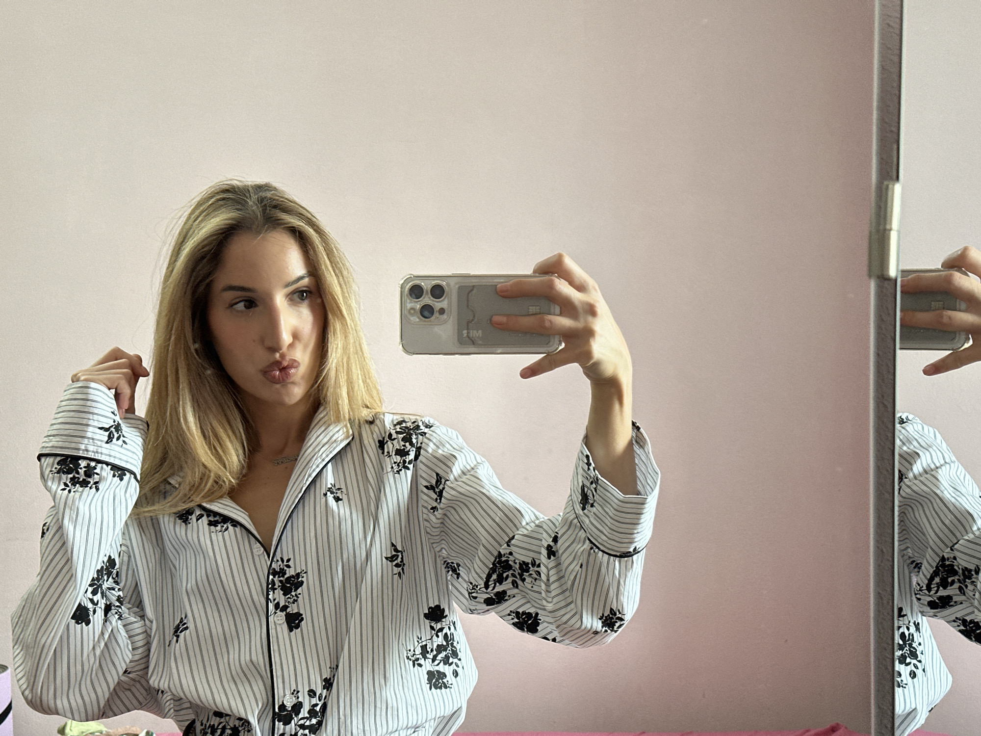A young woman with blonde hair is taking a selfie in a mirror. She is wearing a white pajama set with black pinstripes and a floral pattern. She has her lips puckered and is holding her phone in her right hand, capturing her reflection. The background is a plain pink wall. The mirror is tall and has a metallic frame.