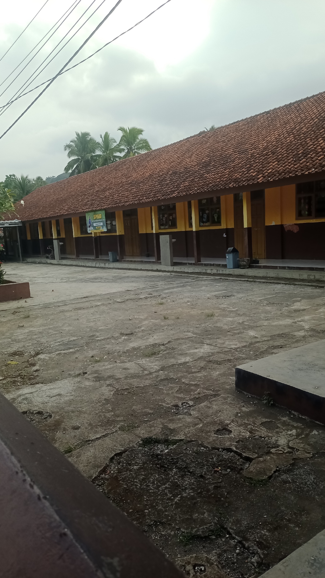 The image captures an outdoor scene of a school building with a red-tiled roof and yellow and brown walls, likely located in Karyamandala, Indonesia. In front of the building is a paved courtyard with some cracks and patches of dirt and vegetation. The courtyard appears empty of people. The building has several windows and doorways, and some posters are visible on the exterior walls. A few trees, possibly palm trees, are seen behind the building. Overhead, power lines crisscross the sky, which is overcast with a light gray color. To the right of the building, a blue trash bin is visible. A dark brown railing or structure is in the foreground on the left side of the image, partially obscuring the view of the courtyard.