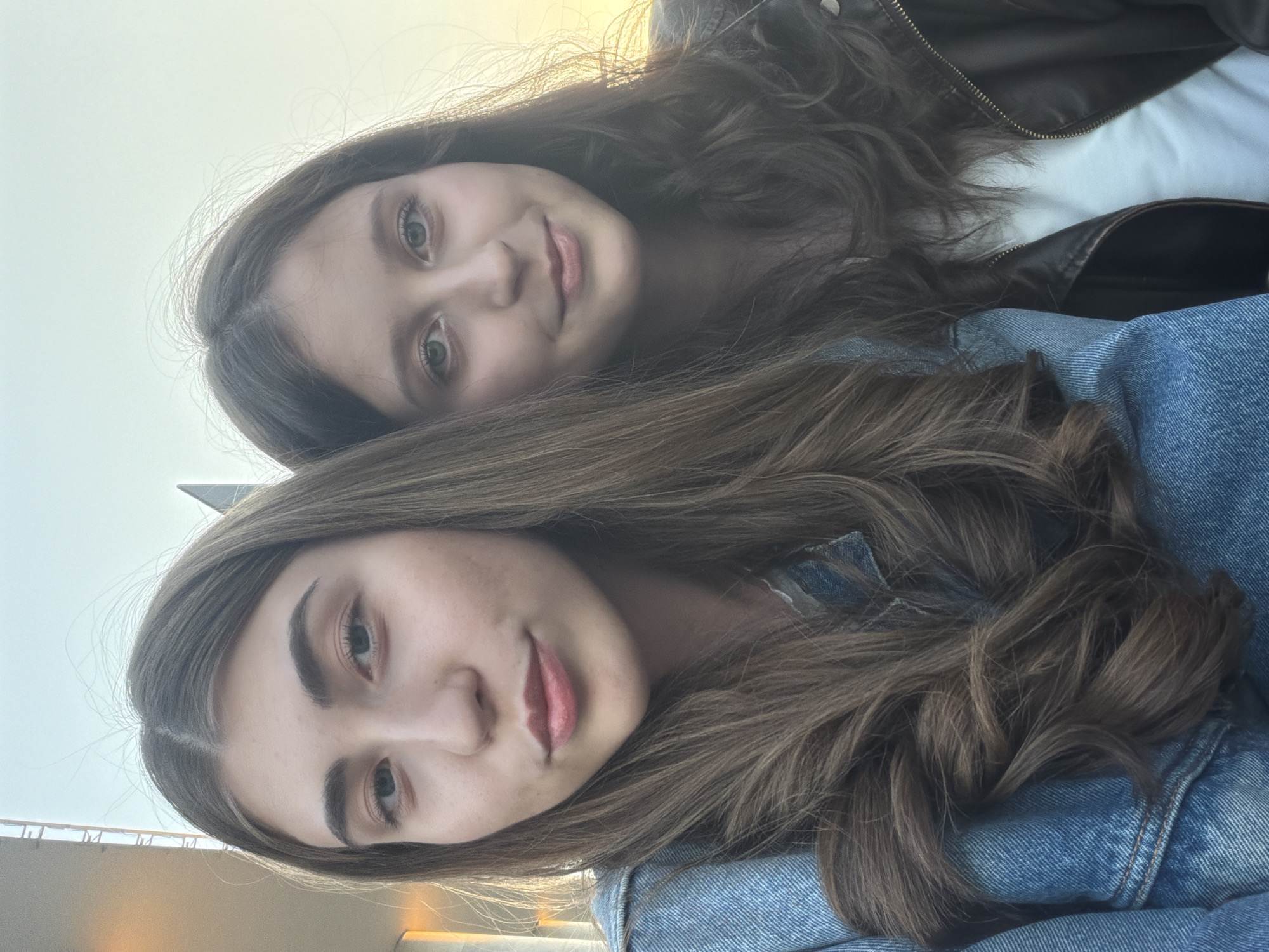Two young women are posing closely together, likely taking a selfie. The woman in the foreground has long, wavy brown hair, a light complexion, and is wearing a blue denim jacket. The woman behind her has straight, dark brown hair, a similar light complexion, and is wearing a white shirt with a brown leather jacket. The background is blurry, but appears to be a slightly overcast sky. It is not possible to confirm the location of the photo is in Krasnoyarsk, Russia from this information.
