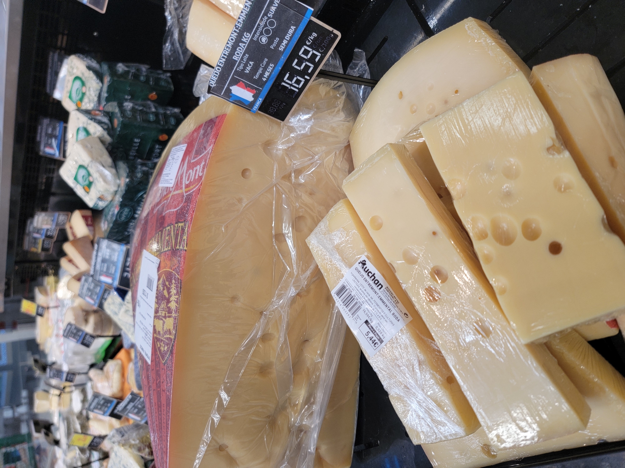 The image shows a display of cheeses in a retail environment, likely a supermarket or cheese shop. In the foreground, several blocks of yellow cheese, some with visible holes, are wrapped in plastic. A large, round wheel of cheese, also wrapped in plastic, is prominent in the background. A price tag with "16,59" is visible, indicating the price per kilogram for a cheese labeled "QUEIJO ENTRETOMONTEMMMENTAL RODA". The lighting suggests an indoor setting with artificial light. The overall scene is organized, with products displayed on shelves. The image does not provide any specific information about the city of Faro, Portugal, nor does it depict any people or actions.