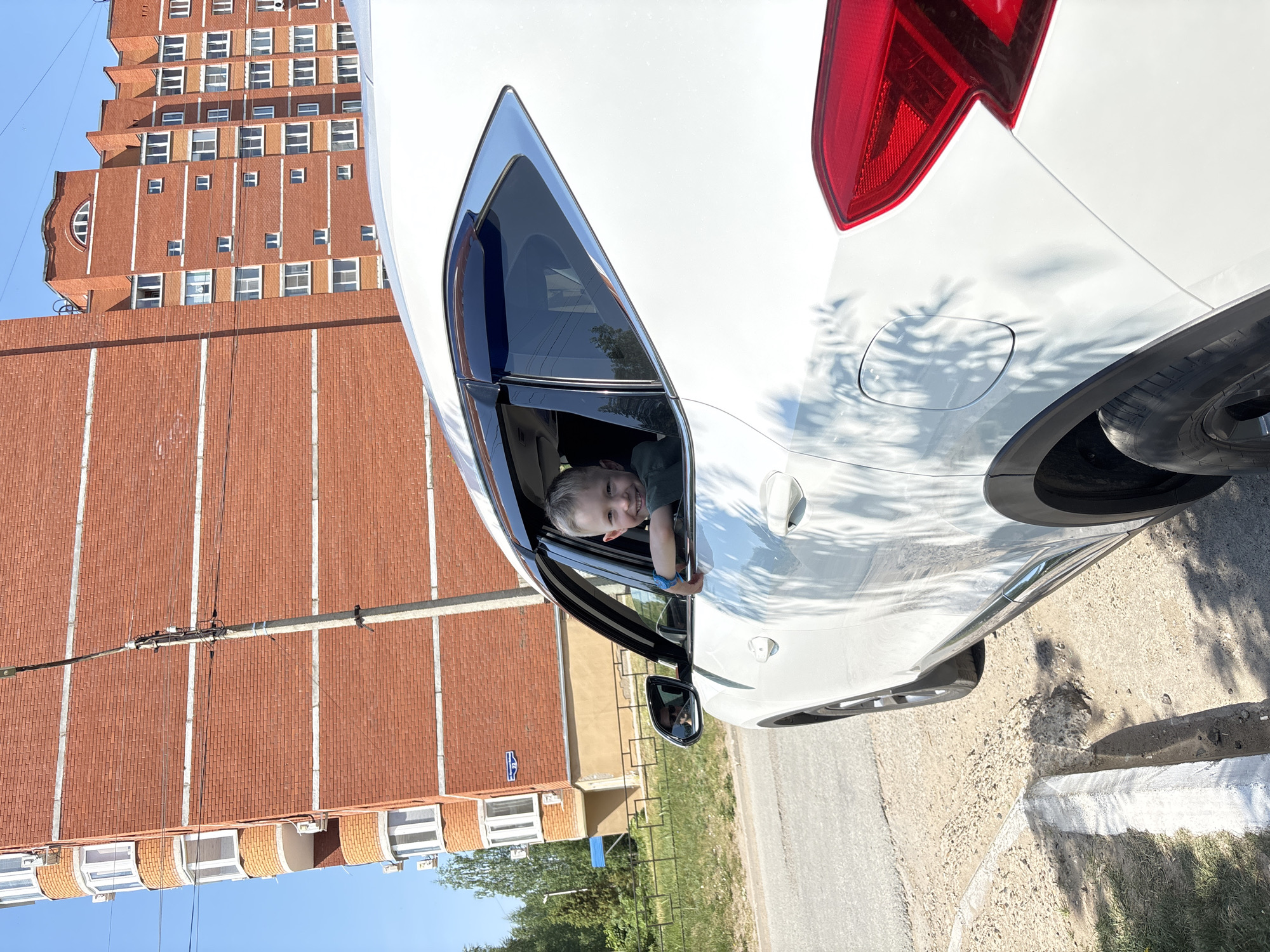 A young child is leaning out of the open back window of a white car. The car is parked on a street in Chaikovsky, Russia. A tall brick apartment building is visible in the background. The street is paved and has a curb. There is a small patch of grass near the curb. The sky is blue.