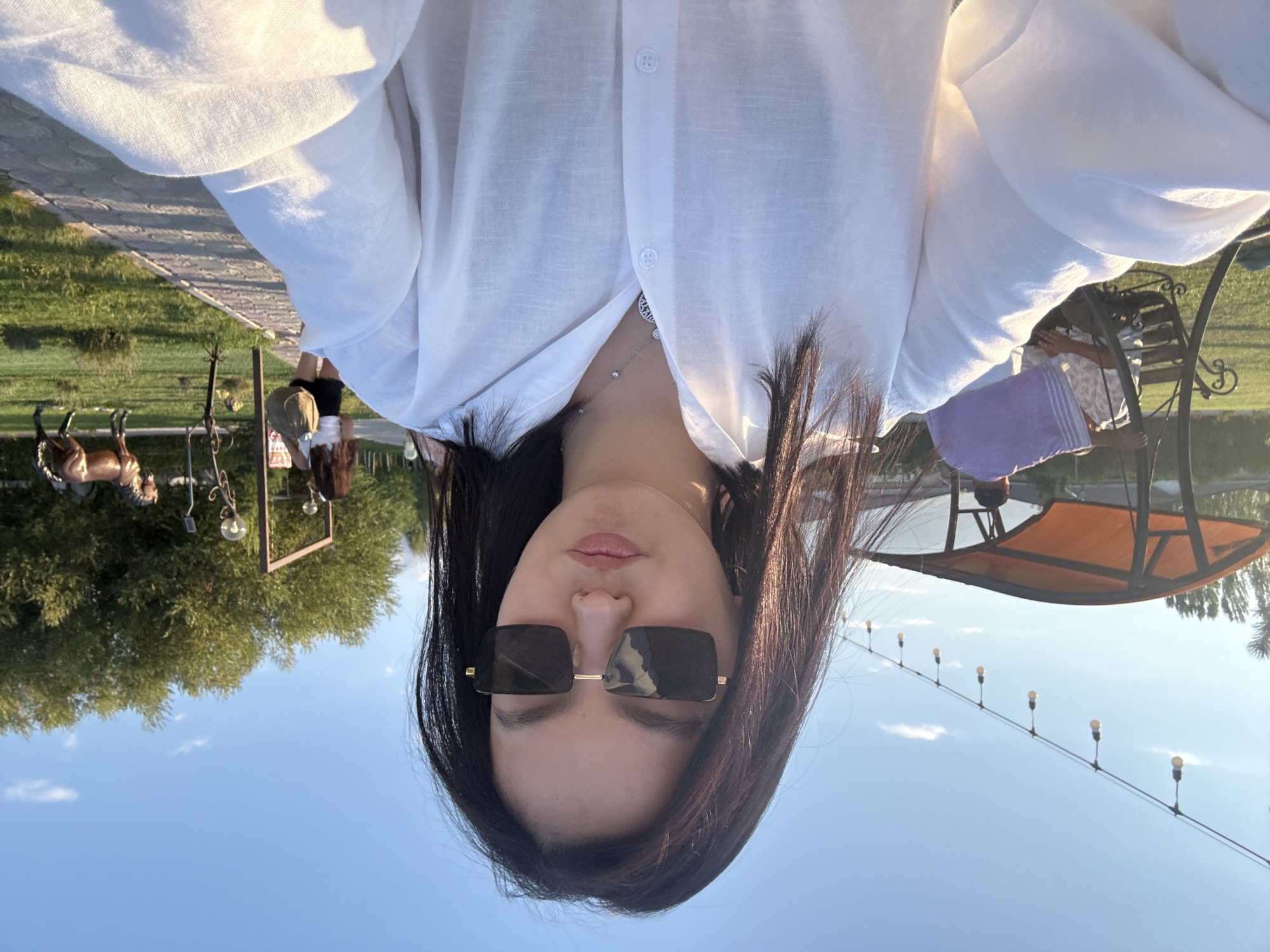 The image captures a selfie of a young woman with long, dark hair, wearing large, square-shaped sunglasses and a white button-down shirt. She is outdoors, with a bright blue sky and scattered clouds above. The setting appears to be a park or outdoor recreational area in Sary-Oy, Kyrgyzstan, suggested by the presence of trees, grassy areas, a paved walkway, and decorative lighting stringed across the background. In the middle ground, a woman in a white top and black shorts walks away from the camera, carrying a woven bag. Further in the background, a statue of a horse stands in a grassy area. To the left, a covered seating area with an orange canopy is visible, with two individuals seated on a bench underneath. One person is wearing a purple wrap, and another is in a patterned shirt. The overall lighting suggests it is late afternoon or early evening, with the sun casting a warm glow.