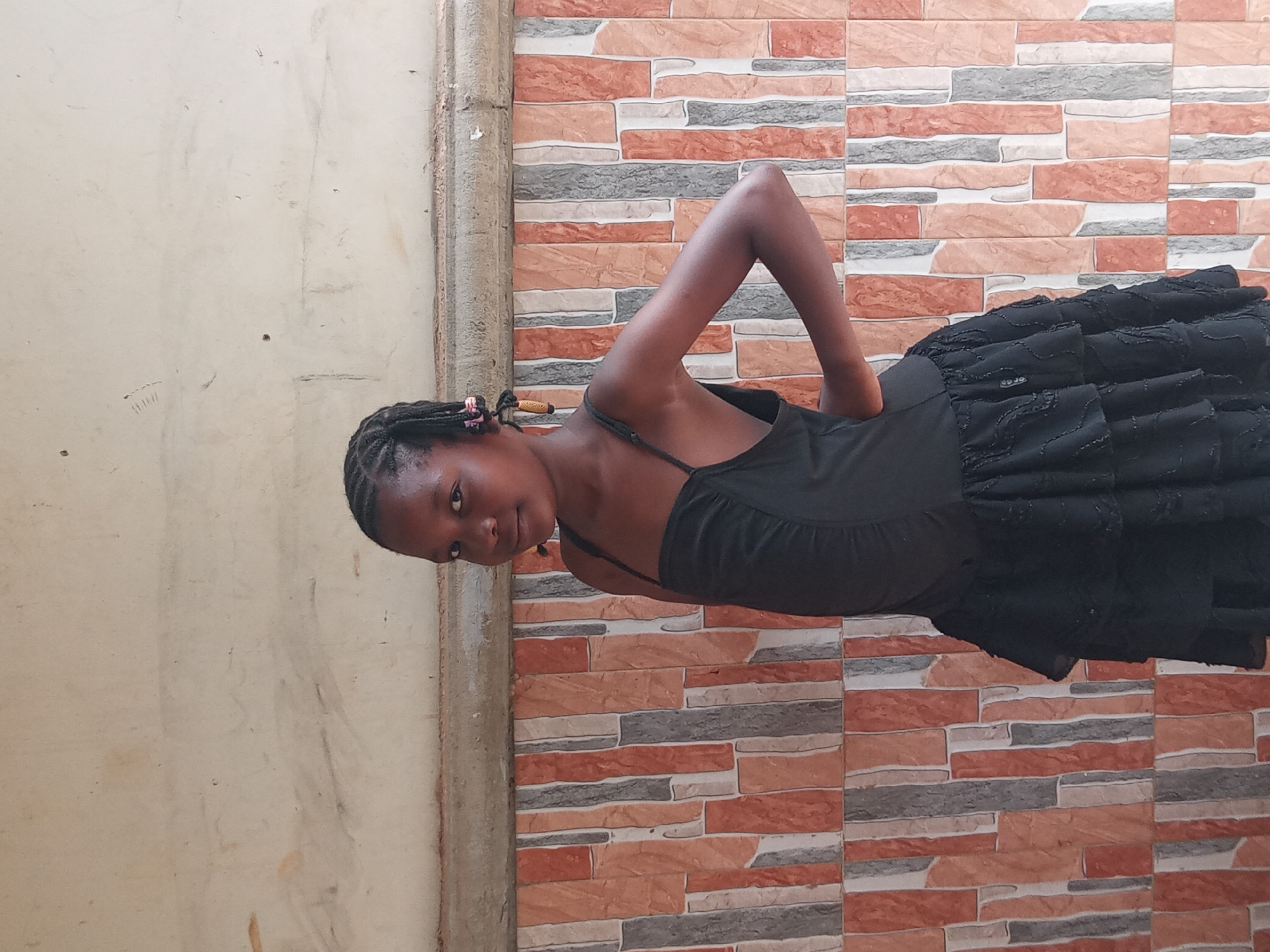 A young Black girl with braided hair, adorned with small wooden beads, is posed against a backdrop of a textured wall with rectangular terracotta and gray tiles. She is wearing a black ruffled dress with spaghetti straps. Her body is angled, with her right arm bent behind her back, and she is looking towards the viewer with a gentle smile. The lighting suggests it is daytime, possibly outdoors, given the natural textures of the wall. The scene appears to be a casual moment, with no specific event indicated. The overall impression is one of youthful pose and simple attire against a textured, possibly outdoor, environment in Sabon Tasha, Nigeria.