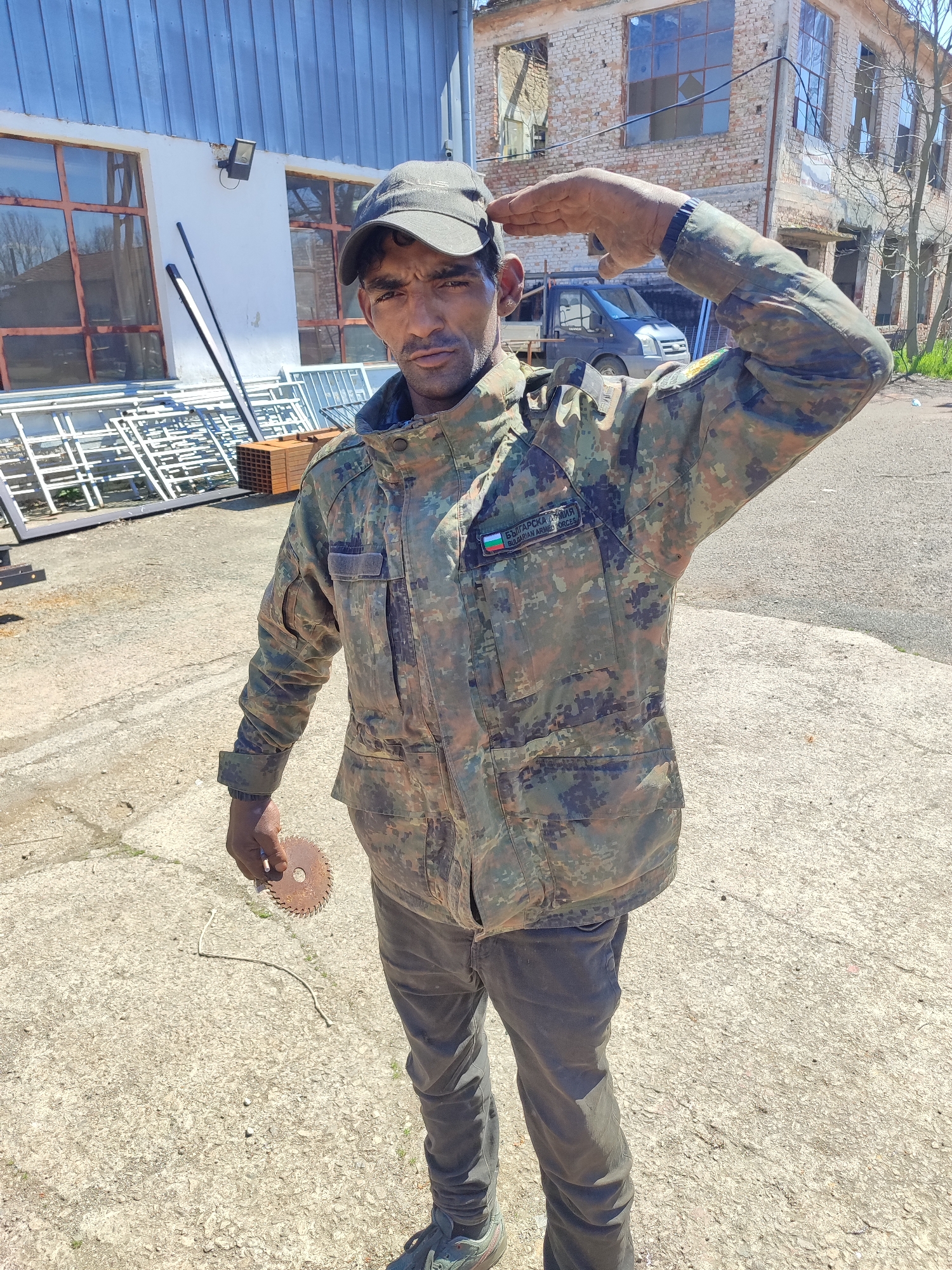 A man in a camouflage military jacket and cap is saluting. He holds a small circular saw blade in his left hand. The jacket has a patch with "Българска армия Bulgarian Armed Forces." The man wears dark pants and athletic shoes. Behind him is a white building with steel framed windows, a pile of steel and wood, a blue van, and a partially destroyed brick building. The ground is concrete. The photograph was taken during the day in bright lighting. It is difficult to confirm the location is Burgas, Bulgaria.